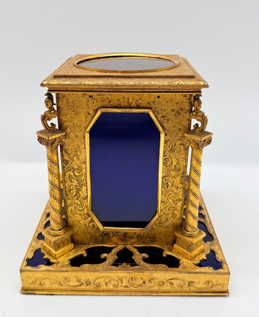 FRENCH GILT-BRONZE AND COBALT GLASS CASKET: An architectural gilt-bronze casket resting on a stepped rectangular base, richly hand-engraved with scrolling foliate arabesques. The central structure features inset bevel-cut deep cobalt glass pane
