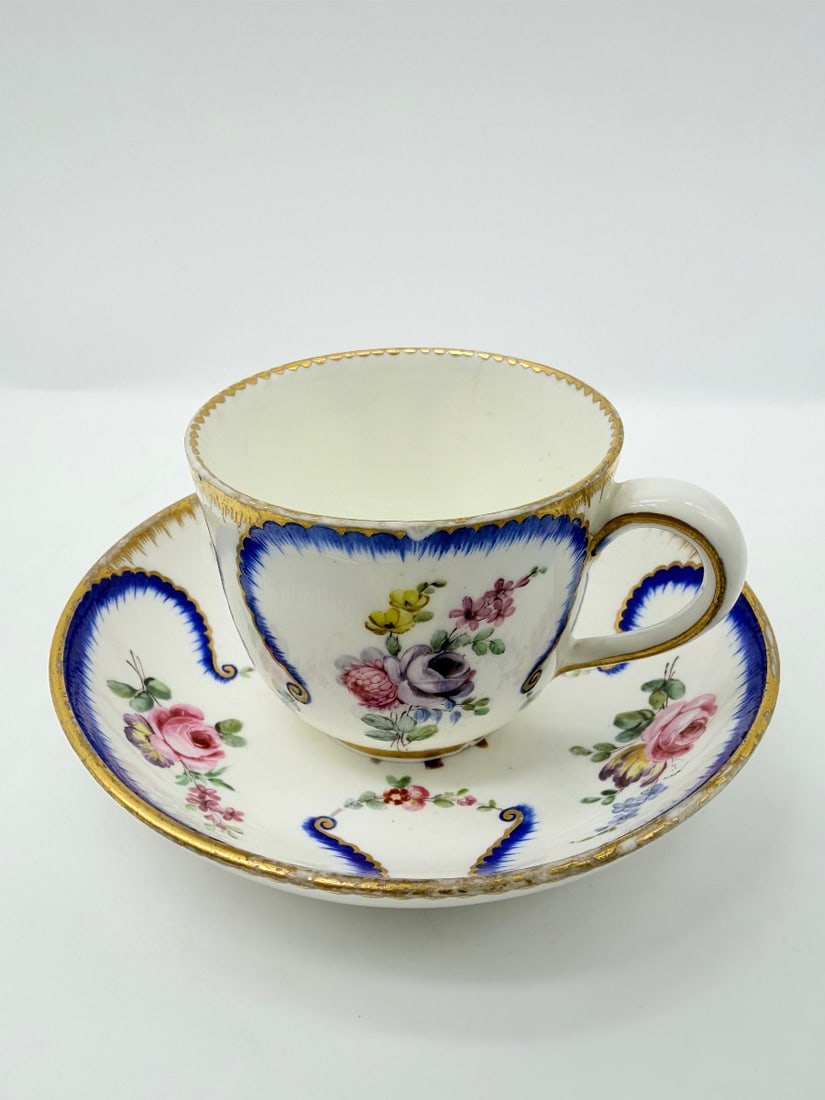 SÈVRES PORCELAIN "FEUILLES DE CHOUX" CUP AND SAUCER: A soft-paste porcelain cup and saucer decorated in the feuilles de choux (cabbage leaf) pattern. Features feathered blue borders edged in gilding, enclosing white reserves painted with polychrome flor