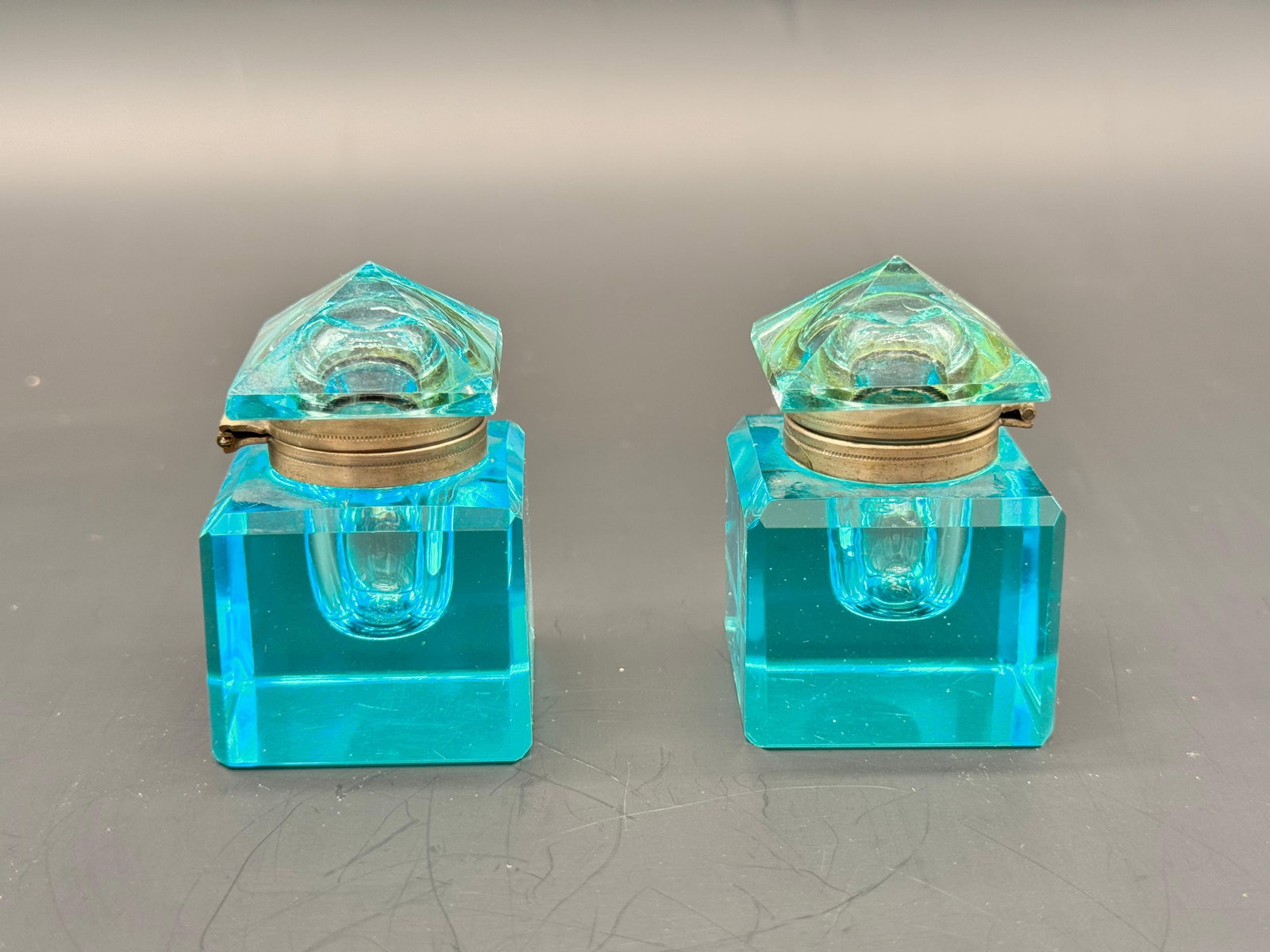 PAIR OF BOHEMIAN AQUA BLUE GLASS INKWELLS: A pair of heavy, cube-shaped Bohemian glass inkwells in a vibrant aqua turquoise color, fitted with hinged brass collars. Each piece is topped with a matching faceted pyramid glass cover attached to t