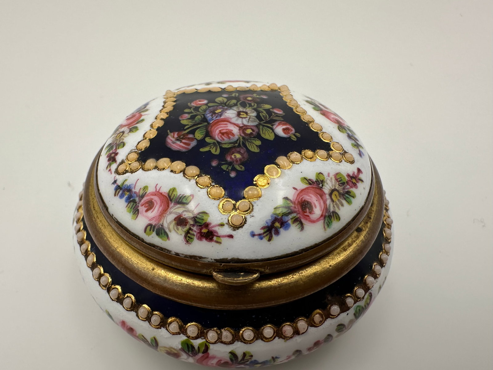 19TH-CENTURY SWISS HAND-PAINTED ENAMEL SNUFF BOX: An antique round Geneva-style enamel snuff or trinket box mounted in gilt metal. The domed lid and body feature a deep cobalt blue ground surrounded by white enamel borders, delicately hand-painted wi