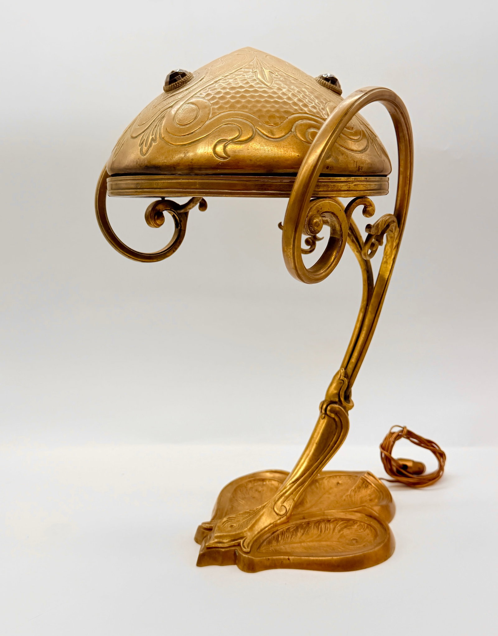ART NOUVEAU GILT BRASS DESK LAMP WITH CABOCHON ACCENTS: An antique Art Nouveau gilded brass desk lamp featuring an organic stem resting on a trefoil organic base. The domed brass shade is decorated with embossed whiplash scrollwork, integrated side loop ha