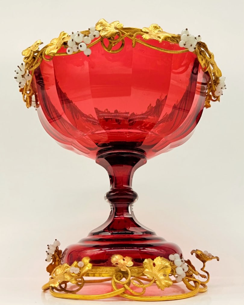 19TH-CENTURY BOHEMIA RUBY CRYSTAL TAZZA WITH GILT BRONZE AND ENAMEL MOUNTS: Footed centerpiece dish (tazza) in deep ruby cut glass or crystal, featuring a wide faceted basin resting on a knopped stem and spreading circular base. Mounted with an elaborate gilt bronze (ormolu) 