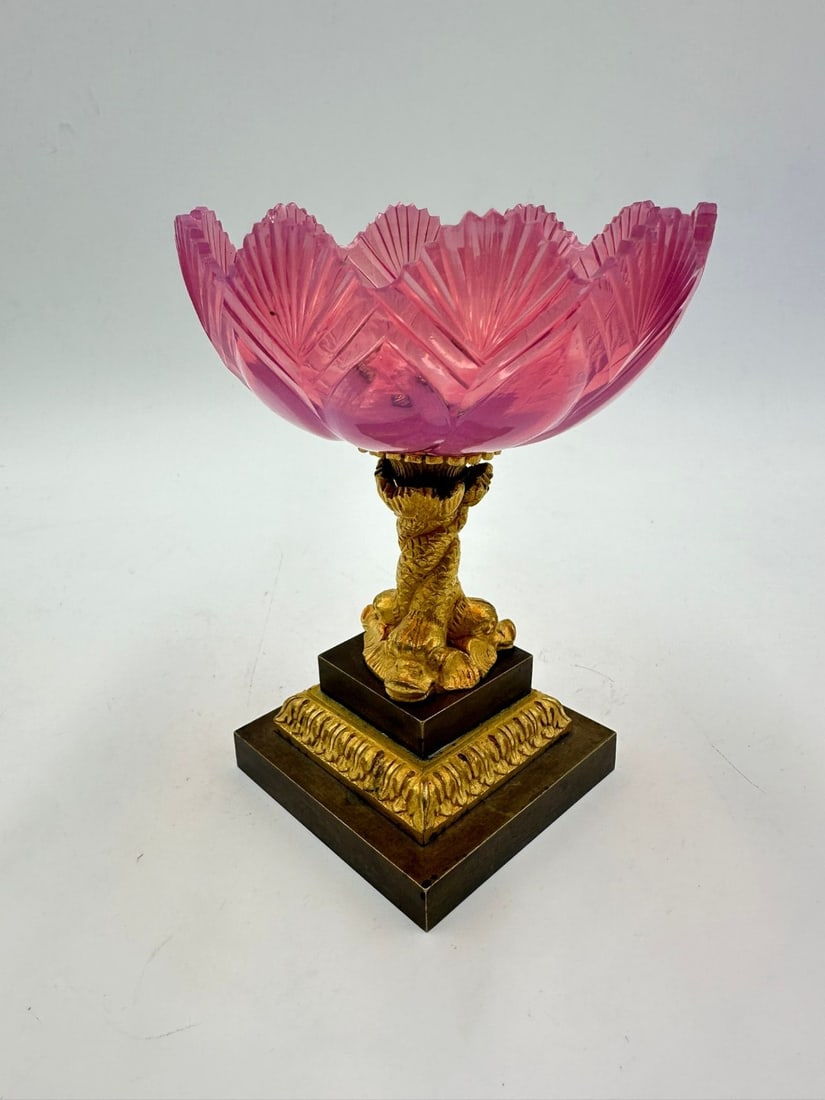 FRENCH GILT-BRONZE MOUNTED CUT GLASS FOOTED TAZZA, CHARLES X PERIOD, CIRCA 1820–1830: A footed tazza or compote dish featuring a pink-tinted cut glass bowl in the pigeon-breast (gorge de pigeon) hue. The bowl is carved with fan and fluted leaf panels along its scalloped rim. It rests o