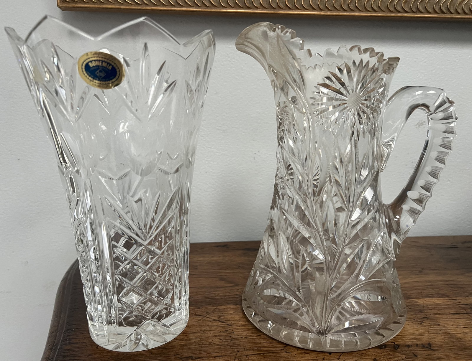 Collection of 2 Cut Glass Pieces a Pitcher and a Bohemia Vase: The image shows two pieces of clear, cut glass.Vase: The piece on the left is a tall vase with a serrated rim. It has a blue and gold sticker that says "Bohemia" on the upper part. The body of the vas