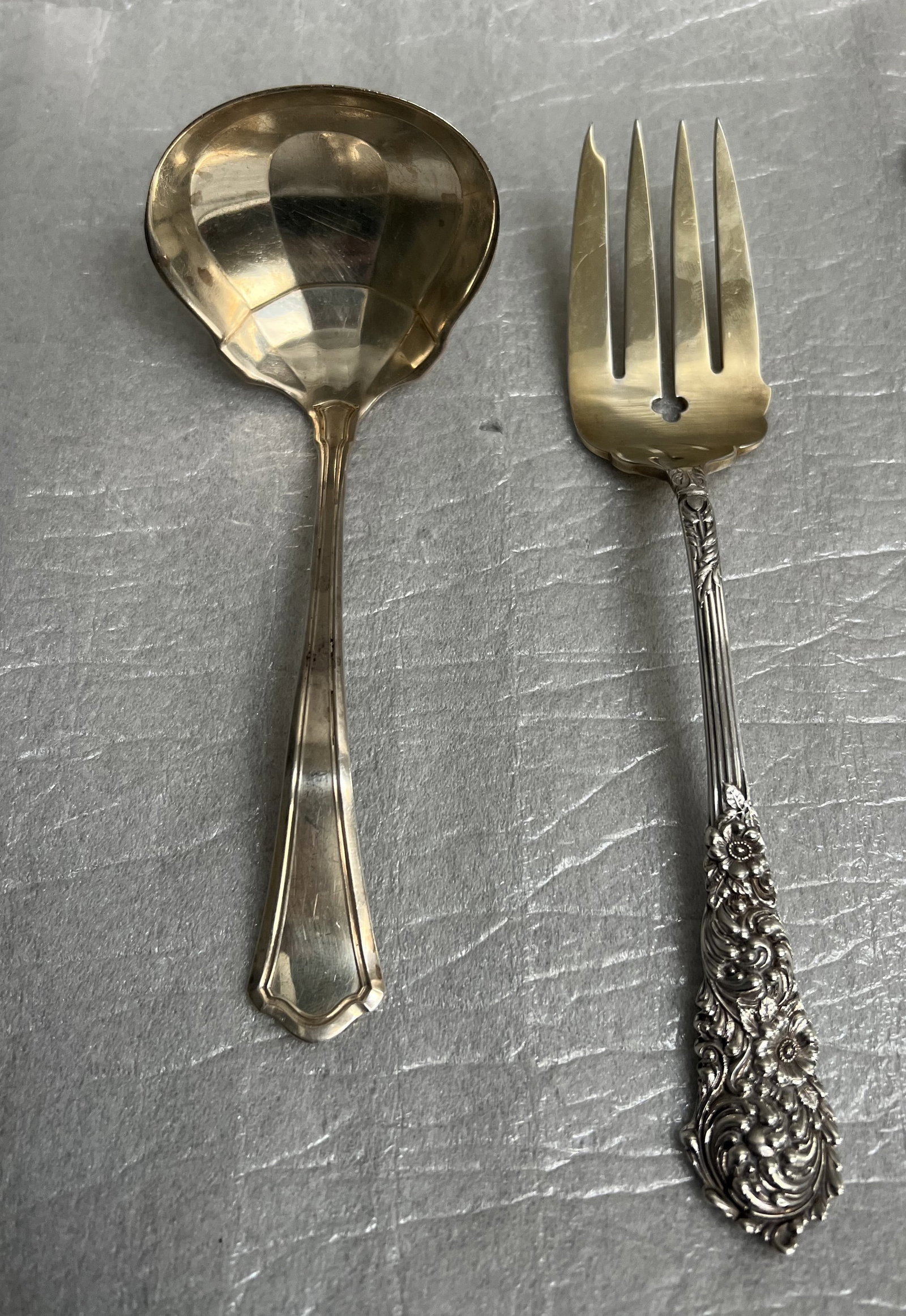 Sterling Silver Gravy Ladle and Serving Fork: Gravy Ladle (Left): This piece has a simple, polished handle with a subtle, elegant outline that flares slightly at the tip. It features a traditional, rounded, bowl-shaped end, which is characteristi