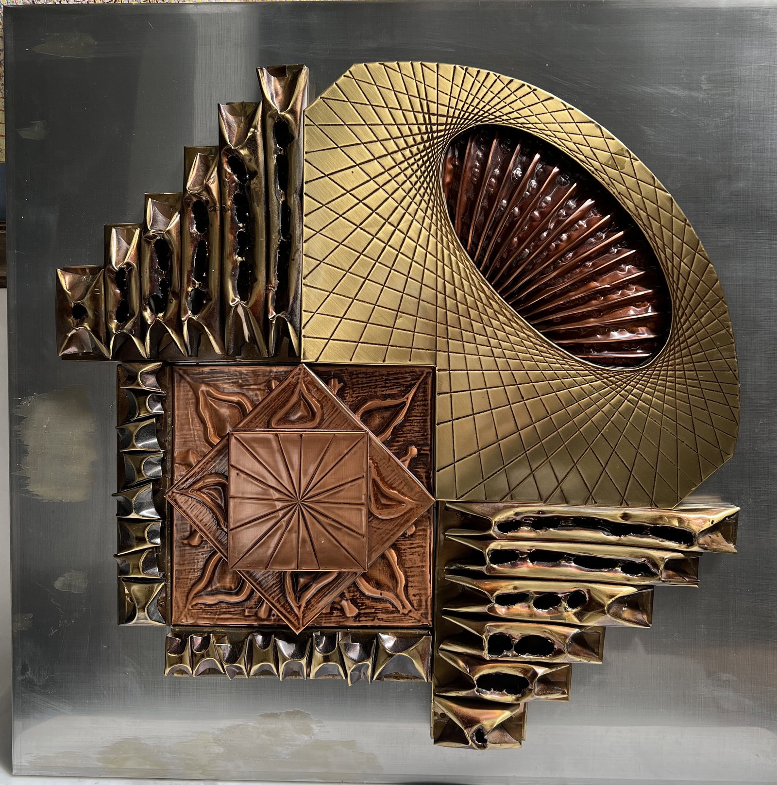Geometric Fusion: Style: Brutalist / Mid-Century Modern Mixed Metal Sculpture.• Materials: Polished steel backplate with applied brass, copper, and torch-cut "burnished" metal elements.• Dimensions: 20" x 20"