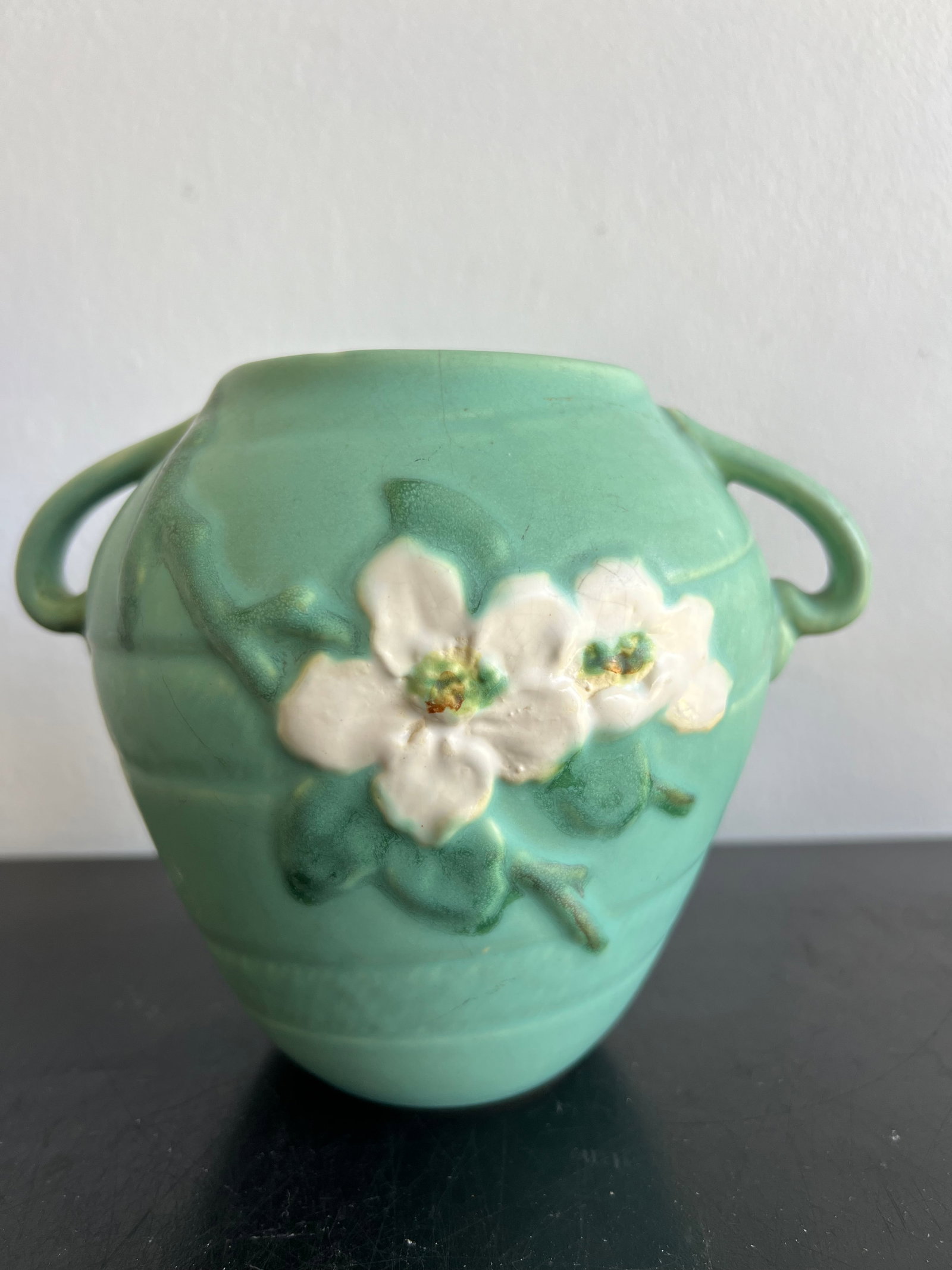 Vintage Weller Pottery Apple Blossom Two-Handled Green Vase (7" H x 8" W): Maker: Weller Pottery (incised signature "Weller" on the base) Pattern / Motif: Apple Blossom; features raised white floral blooms and green leaves on the front against a subtly ridged background Colo