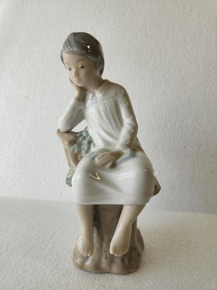 Lladro Thinking Boy with Book on Stump: Lladro, Spain, modeled as a boy seated on a stump, resting his head on his hand in thought, a book open in his lap, the details picked out in delicate shades of grey, beige, and blue, stamped with blu