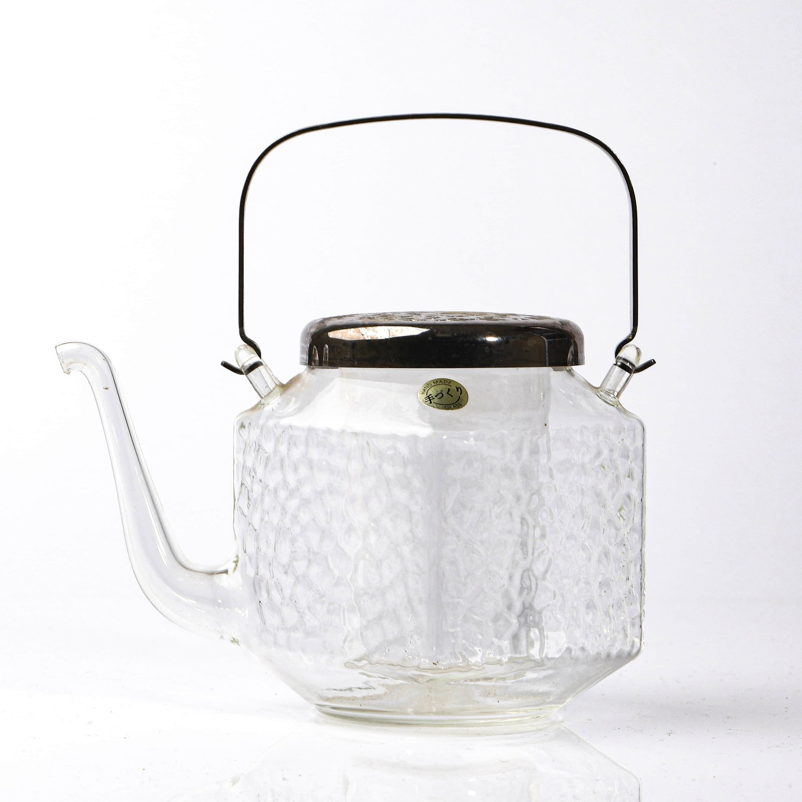 JAPANESE HAND-BLOWN GLASS TEAPOT WITH METAL LID AND HANDLE: Dimensions Length (spout to handle): 6 7/8 in (17.5 cm) Height (to top of lid): 6 1/2 in (16.5 cm) Description A Japanese hand-blown clear glass teapot featuring a hammered-texture body, an elegantly 
