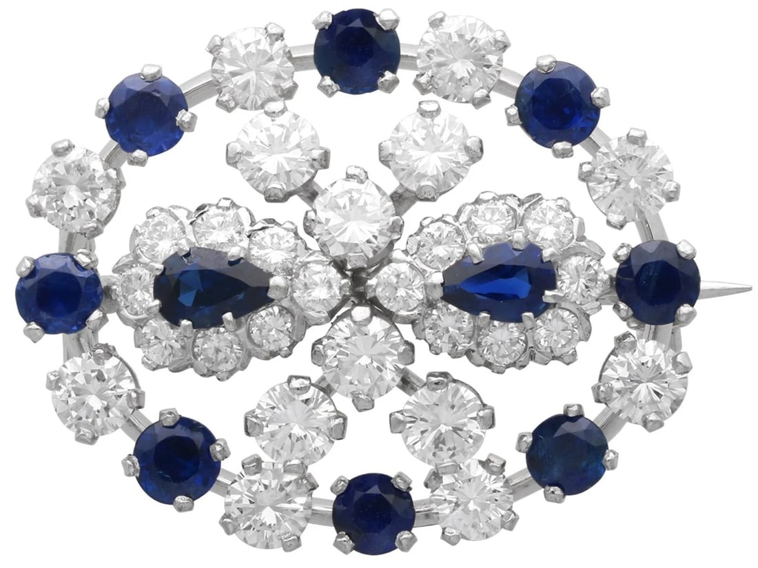 WHITE-METAL BLUE AND COLORLESS STONE OVAL BROOCH: WHITE-METAL BLUE AND COLORLESS STONE OVAL BROOCH Description: An oval openwork brooch in bright white metal, set with alternating deep-blue stones and colorless accents in a symmetrical floral and geo