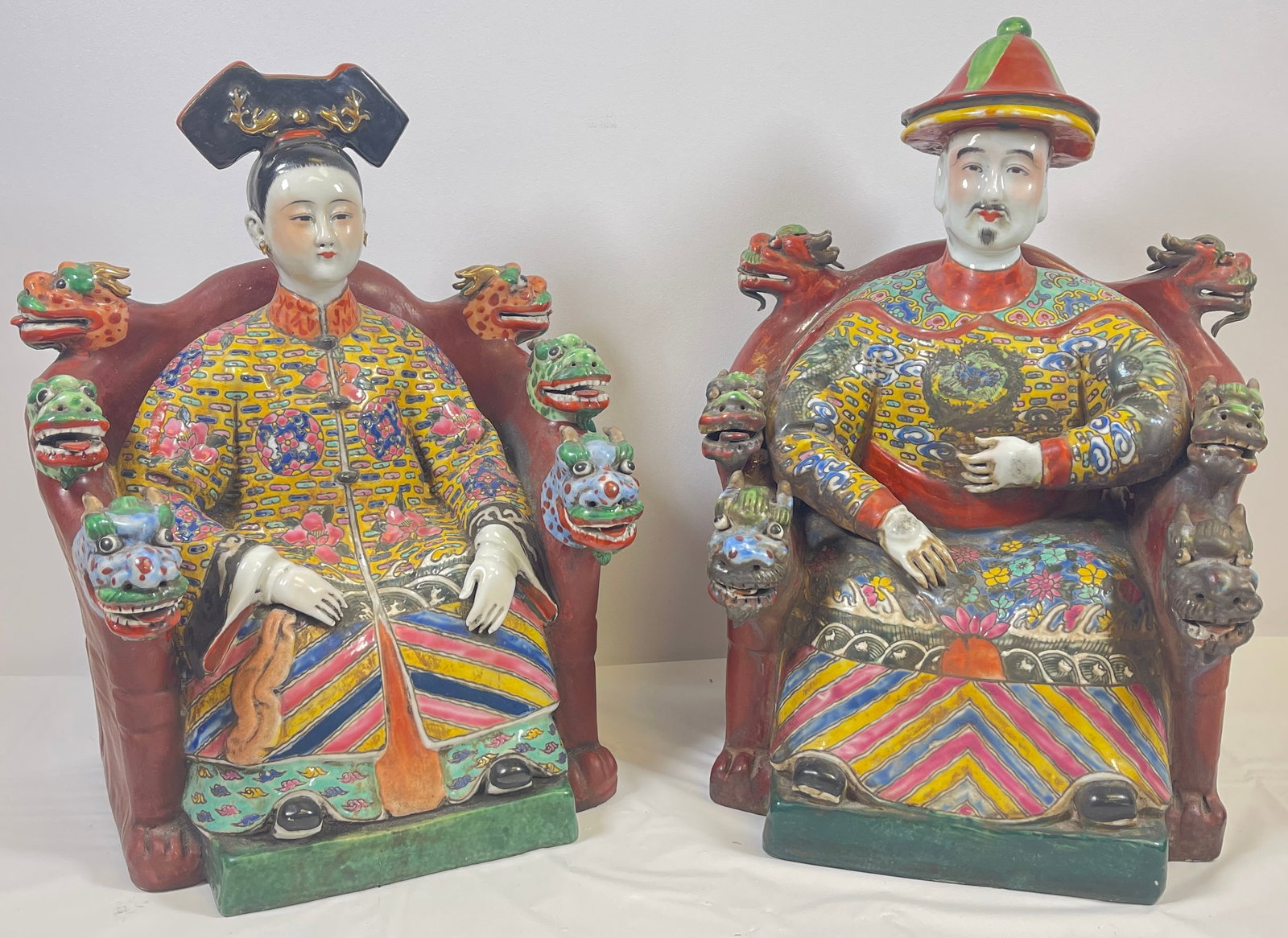 Pair Chinese Porcelain Model of Seated Emporer & Empress: Seated upon red chairs with red, blue and green dragon heads decorating them. The emperor is wearing a red and green hat; both empress and the emperor are dressed in royal attire. Republic period. H: 