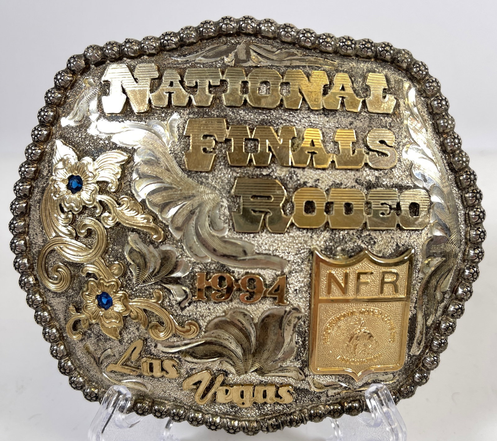 c1994 Sterling Silver & 10K Gold National Finals Rodeo Las Vegas Belt Buckle: Sterling silver buckle with 10k gold filled lettering & adornments, with two sapphires. Dated 1994. Buckle measures 4 inches x 3.5 inches. Numbered 47/500. 4.7 troy ounces (149.1 grams).