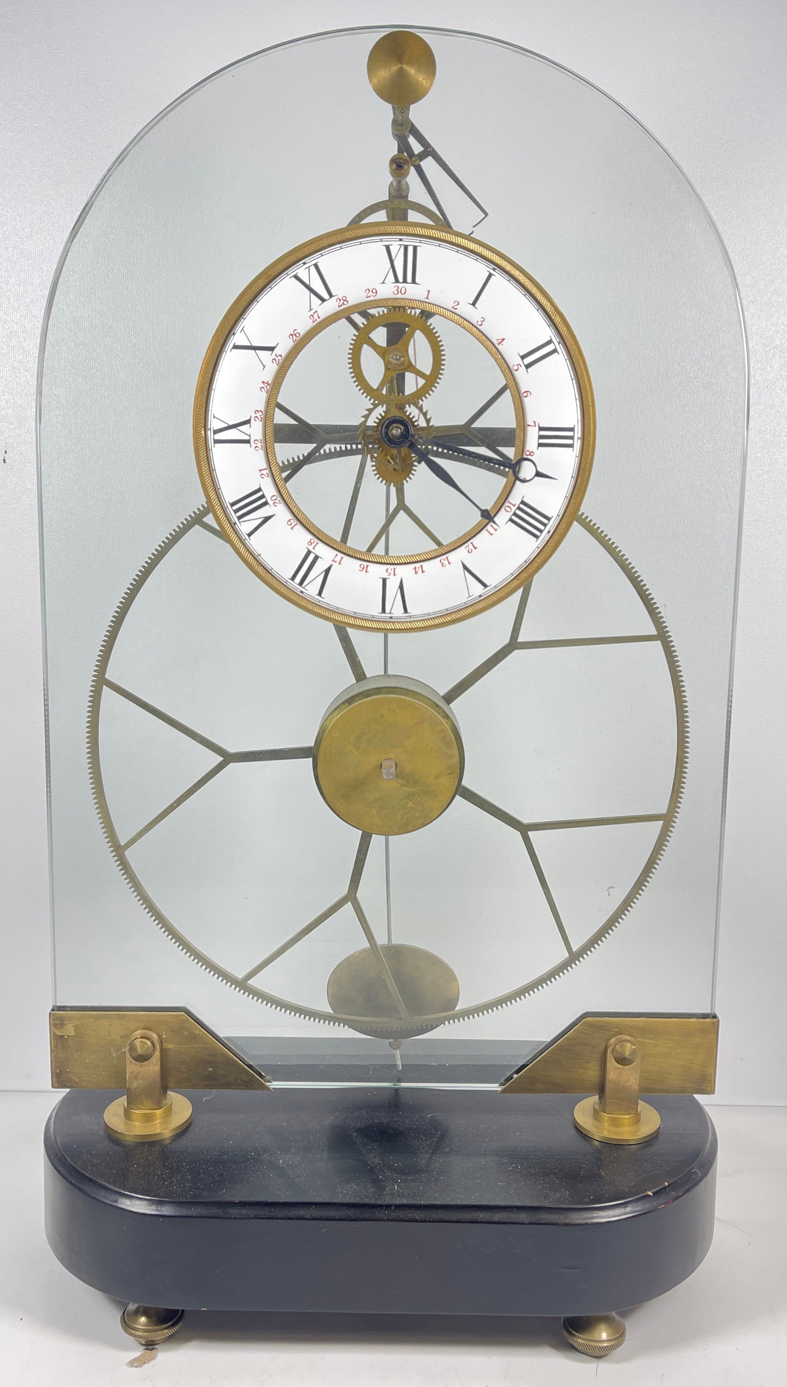 French Great Wheel Skeleton Clock: Exposed brass movement on a arched glass frame and ebonized oval base. Enameled face. Approx. 19.25 inches high x 11 inches wide x 5.75 inches deep. Overall good condition, clock is in running conditi