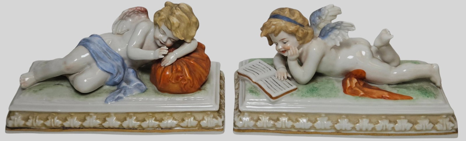 Pair c1900 Meissen Style German Porcelain Cherub Figurines: One depicting a sleeping cherub and the other reading a book.They measure approx 3.25 inches high, 5.75 x 2.75 inches. Overall good condition, no chips or cracks.