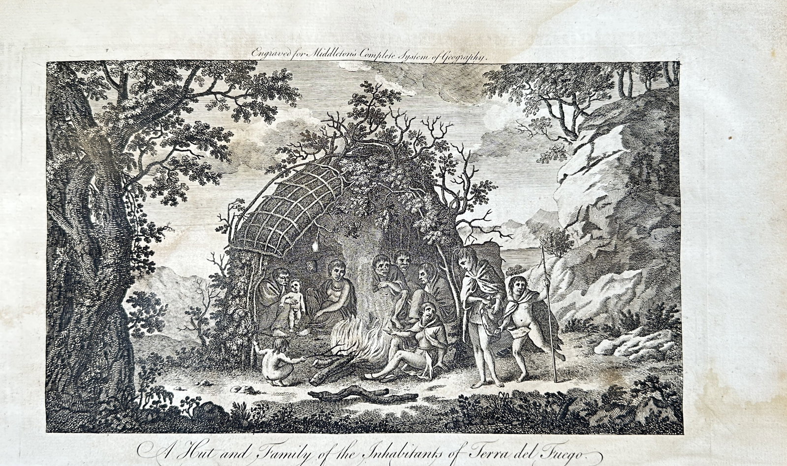 Alexander Buchan – A Hut and Family of the Inhabitants of Tierra del Fuego – Engraving (1779): A detailed engraving after a drawing by Alexander Buchan, depicting a family of the indigenous people of Tierra del Fuego gathered outside a hut made of branches. A man stands with a spear and a small