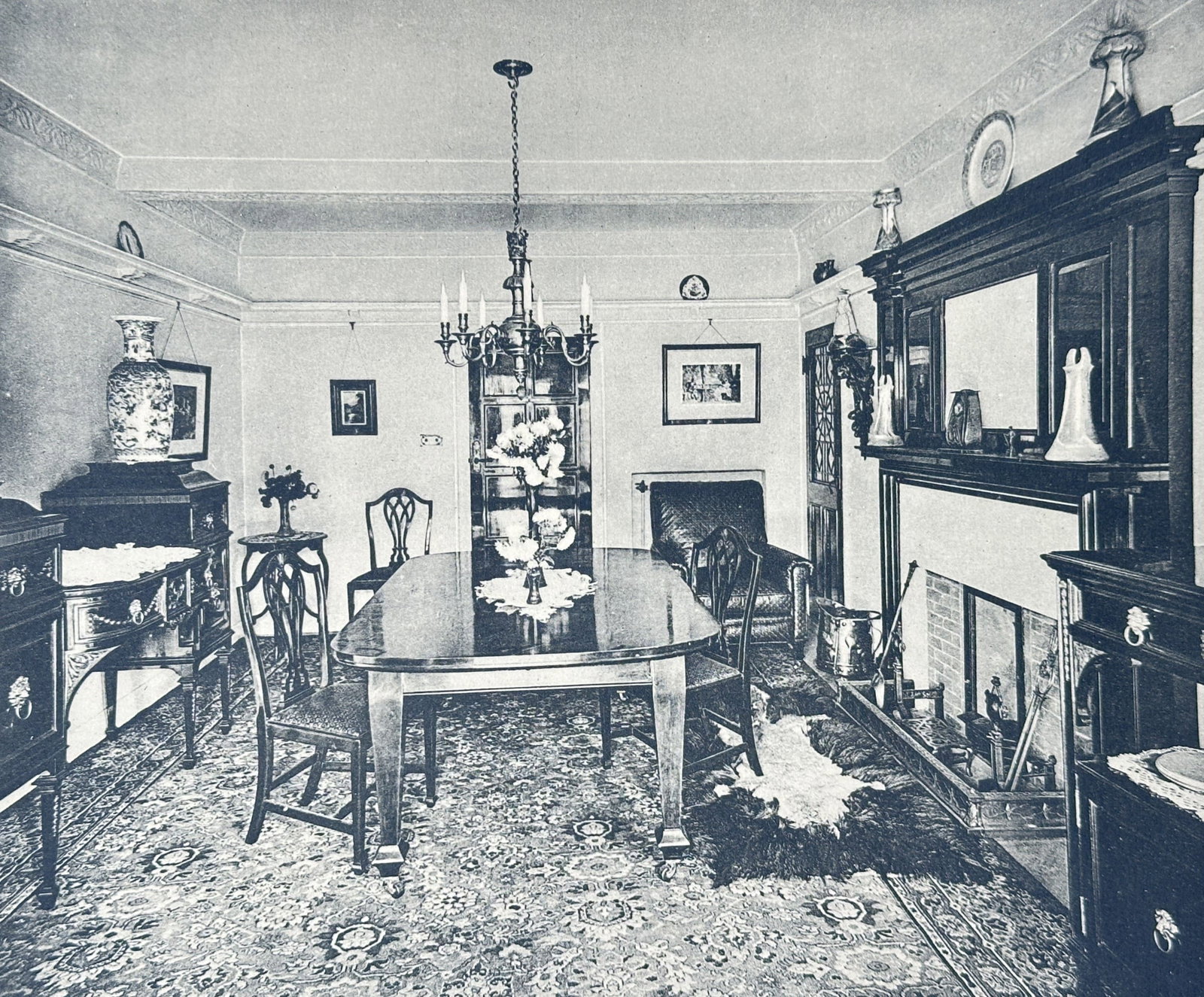 Herbert Arthur Welch – Dining Room at Golders Green Residence – Collotype (1910): "The English Interior" This print features a view of a formal dining room furnished with a large wooden table, matching chairs, an elaborate patterned rug, and a monumental wooden fireplace surround. 