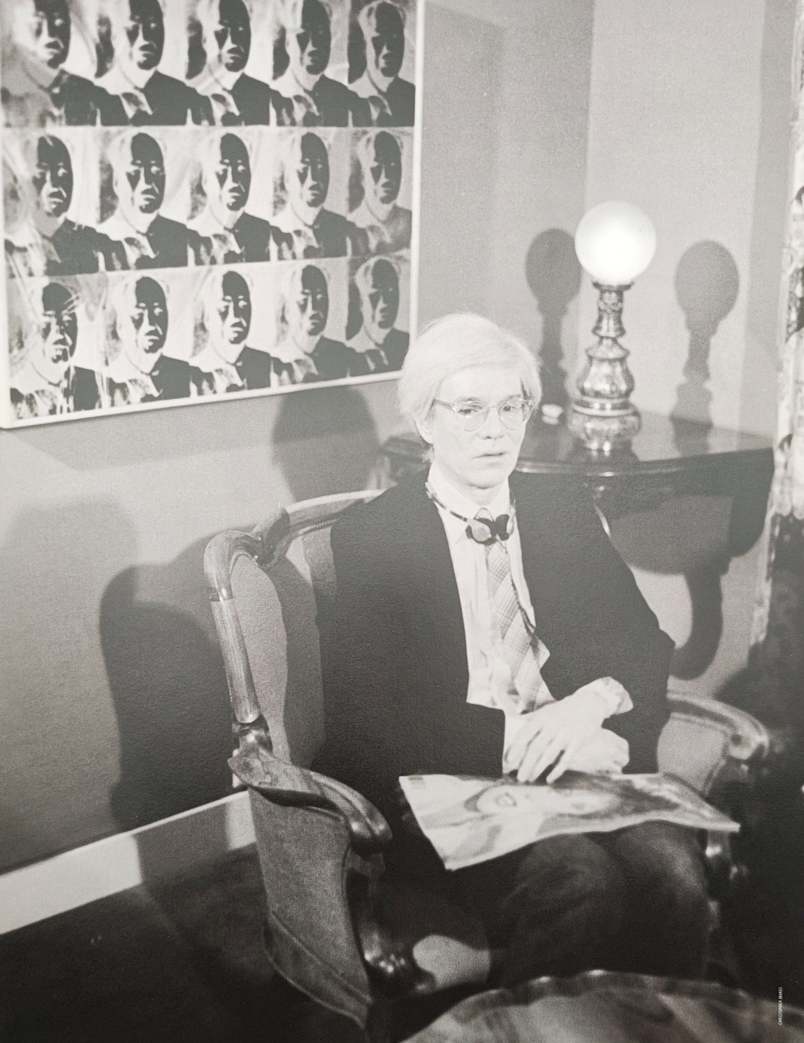 Andy Warhol,after – Andy Warhol Seated with Jackie Portrait – Offset lithographic reproduction: "Andy Warhol Seated with Jackie Portrait" presents an interior photographic portrait depicting the artist resting in an armchair beneath a silkscreened canvas grid of Jacqueline Kennedy. Published in 