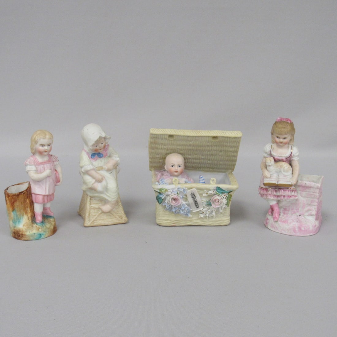 4 HEUBACH FIGURINES:: BABY IN HIGHCHAIR 4.5 IN. H STAMPED, GIRL NEXT TO TREE STUMP, 4.5 IN. STAMPED, GIRL SITTING ON BRICK WALL READING WITH CAT NOT STAMPED & BABY IN A BASKET WITH FLOWERS 4.5 IN. STAMPED DEPOSE. CHIPS ON 