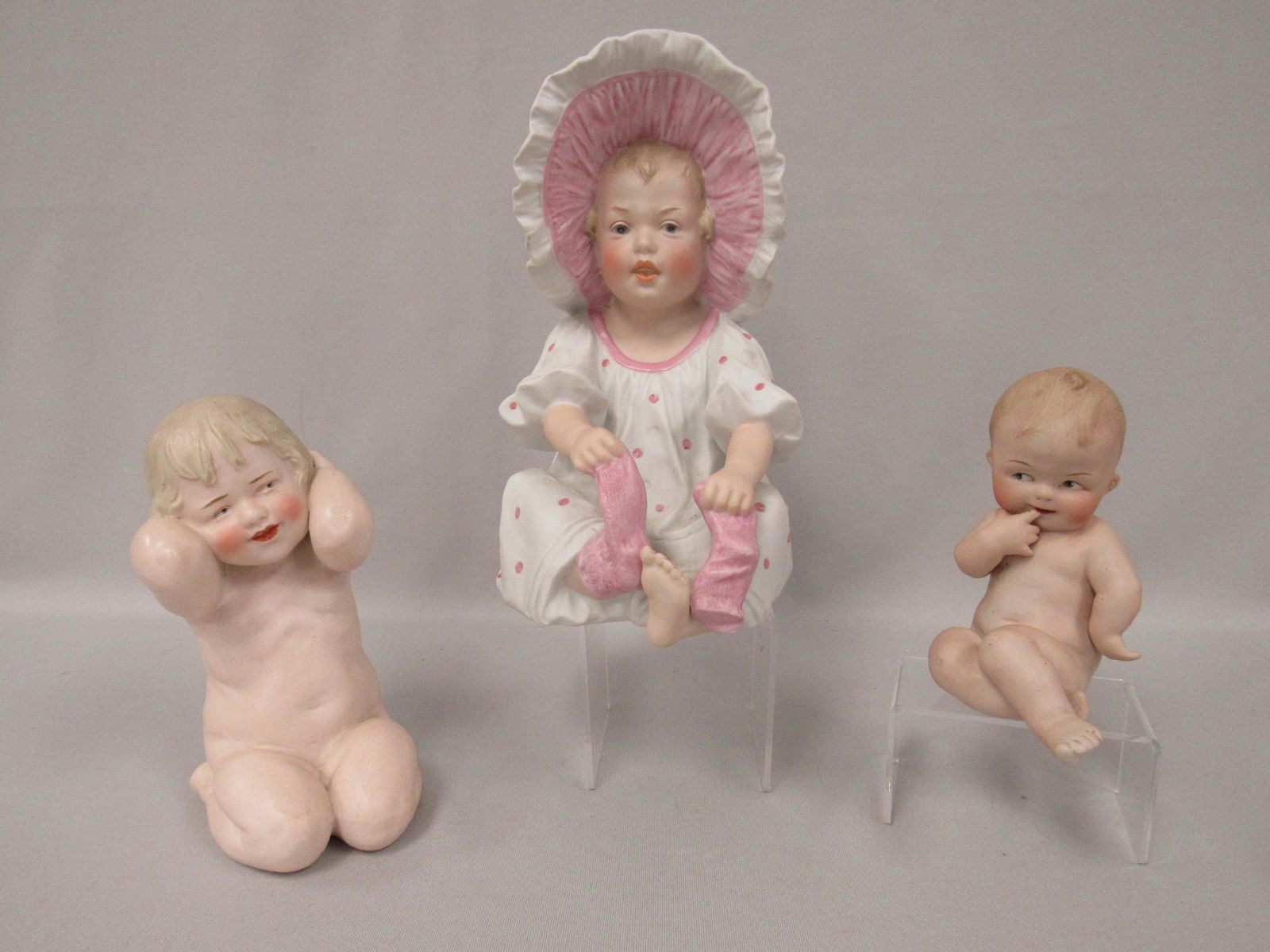 (3 PCS.) GEBRUDER HEUBACH FIGURINES:: 8 IN. BABY GIRL WITH LARGE BONNET PULLING HER SOCKS OFF (SUNBURST MARK) 8 IN. H, 5 IN. W, 6 1/2 IN. D, SMILING CHILD WITH ARMS OVER HIS EARS, 6 1/2 IN., 3 1/2 IN. W, 4 IN. D, & COY BABY WITH GOOGLY