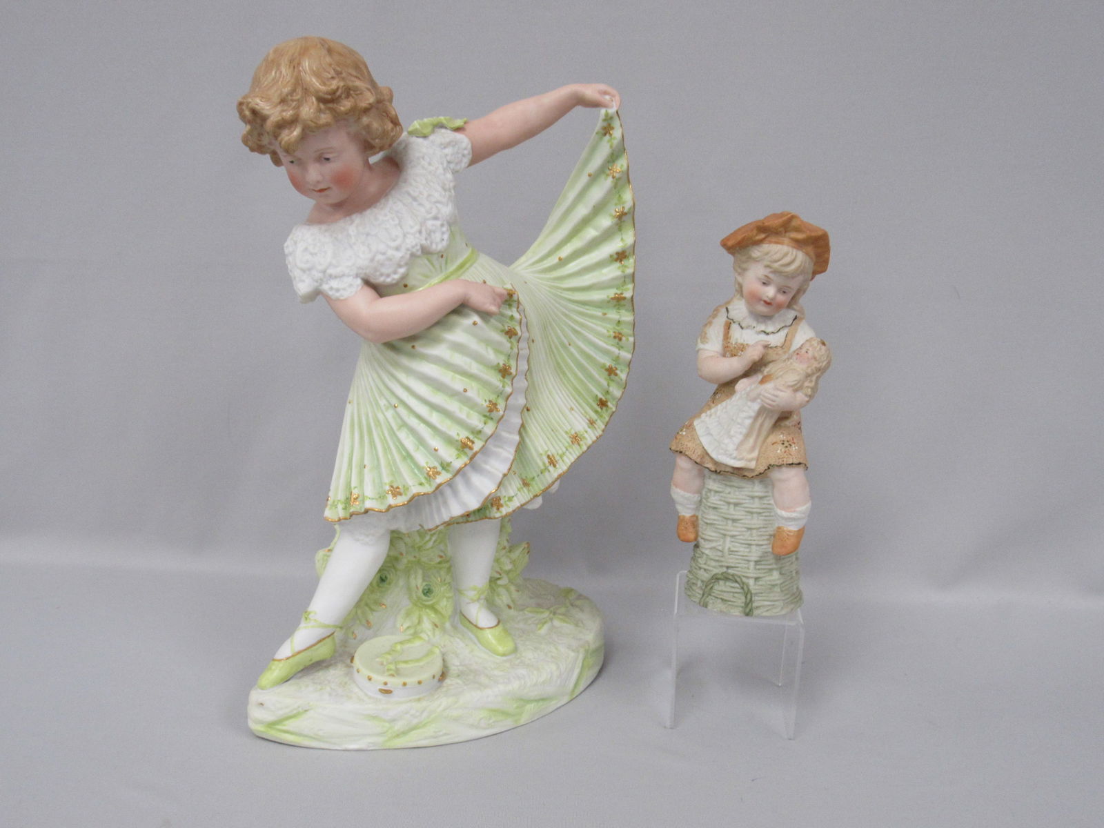 (2 PCS.) HEUBACH DANCING GIRL & GIRL WITH DOLL:: WONDERFUL FINE QUALITY DANCING GIRL CIRCA 1900, POSED MID-DANCE WITH HER ELABORATE PLEATED SKIRT EXTENDED, PALE GREEN DRESS WITH GOLD TRIM. 15 IN. H., 9 1/2 IN. W., 6 IN. D. & HEUBACH FIGURINE OF A