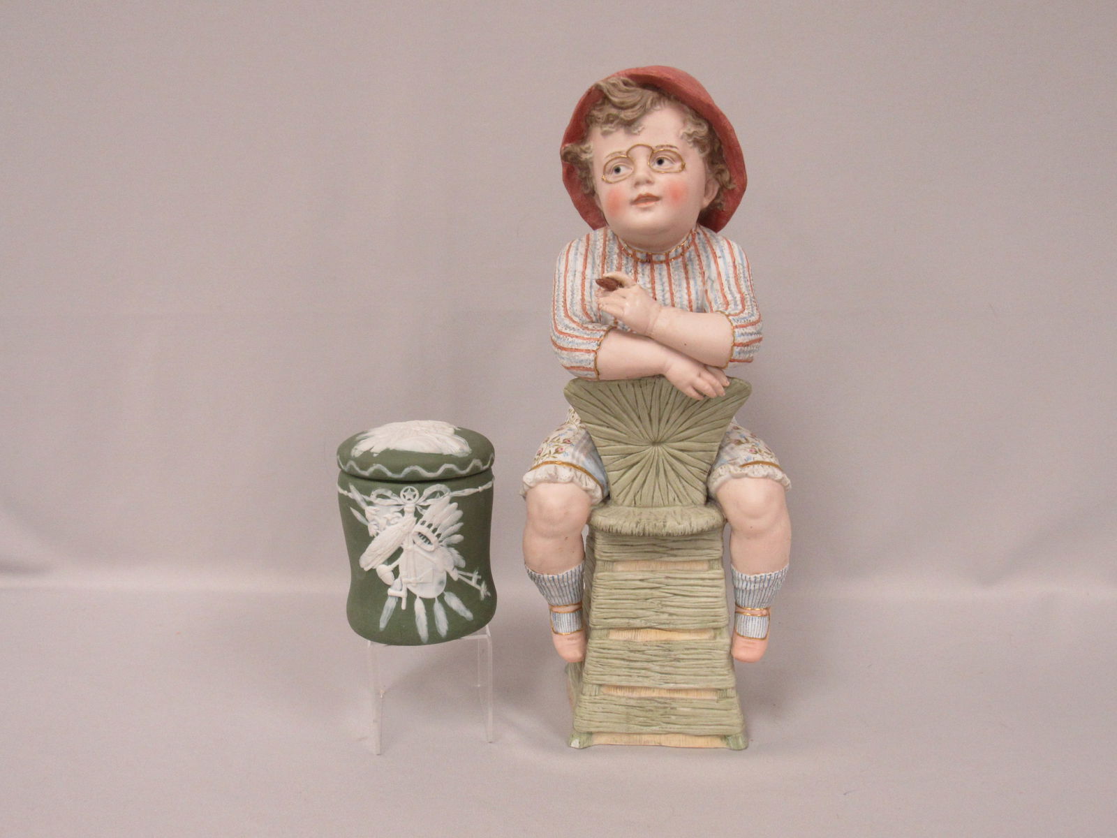 (2 PCS.) HEUBACH BOY WITH CIGAR & TOBACCO JAR:: WONDERFULLY MOLDED & PAINTED BISQUE FIGURE OF A YOUNG BOY ON A WICKER CHAIR HOLDING A CIGAR, TIP OF CIGAR WAS REPAIRED, 17 IN. H, 7 IN. W, 6 IN. D & A JASPERWARE TOBACCO JAR (SUNBURST MARK) FEATURING