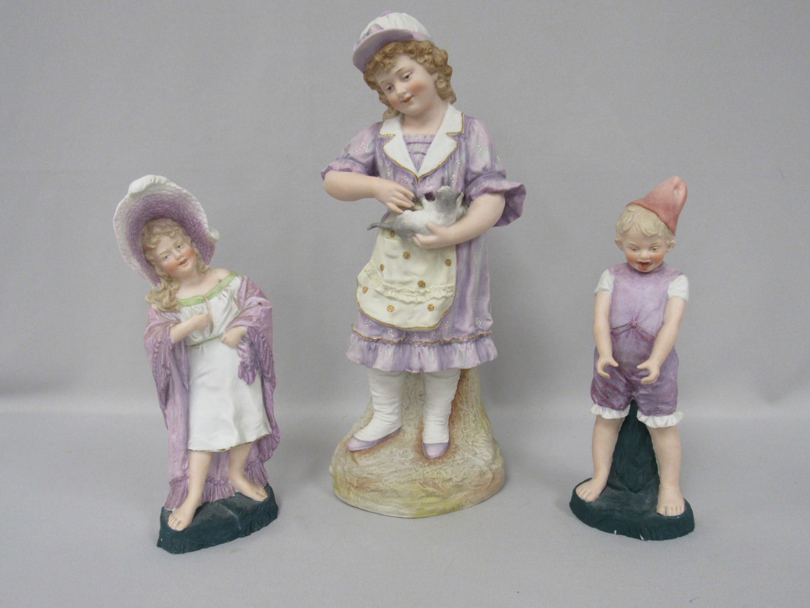 ROUP OF 3 GERMAN BISQUE FIGURINES:: CIRCA 1900, EACH IS ATTRACTIVELY DRESSED IN A LAVENDER PALETTE. EACH FEATURE A YOUNG CHILD. THE LARGEST IS A 14 IN. GIRL HOLDING A CAT, 14 IN. H, 5 1/2 IN. W, 4 IN. D, A 10 IN. H SWEET YOUNG GIRL