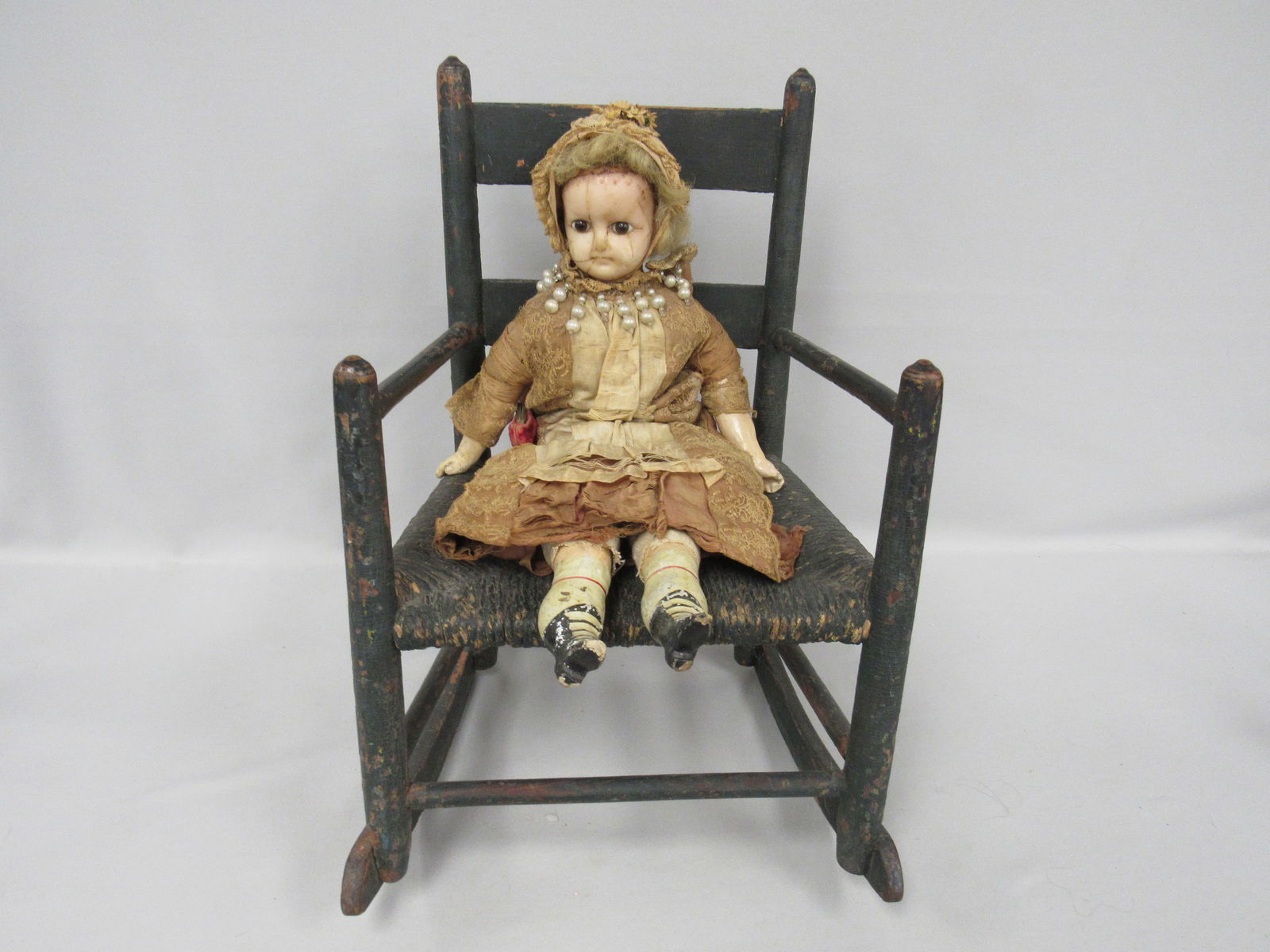 20.5 IN. WAX OVER PAPER MACHE:: SHOULDERPLATE, GLASS EYES. CRACKS & FADING ON FACE & PLATE CLOTH BODY WITH COMPOSITION ARMS & LEGS (REPAIRS TO LEGS) VERY FRAGILE DRESS NOT REMOVED. IN OLD PAINTED ROCKER (SOME CHIPPING, TOE CHIPS),