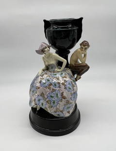 A Goldscheider Art Deco porcelain group figurine of a faun with a young lady.: Girl with Faun sitting on a Vase. Girl wearing a cavalier hat and a colorful wide skirted dress. The Satyr leans on a green aquamarine vase, both are on a black plinth. Marked Goldscheider and artist
