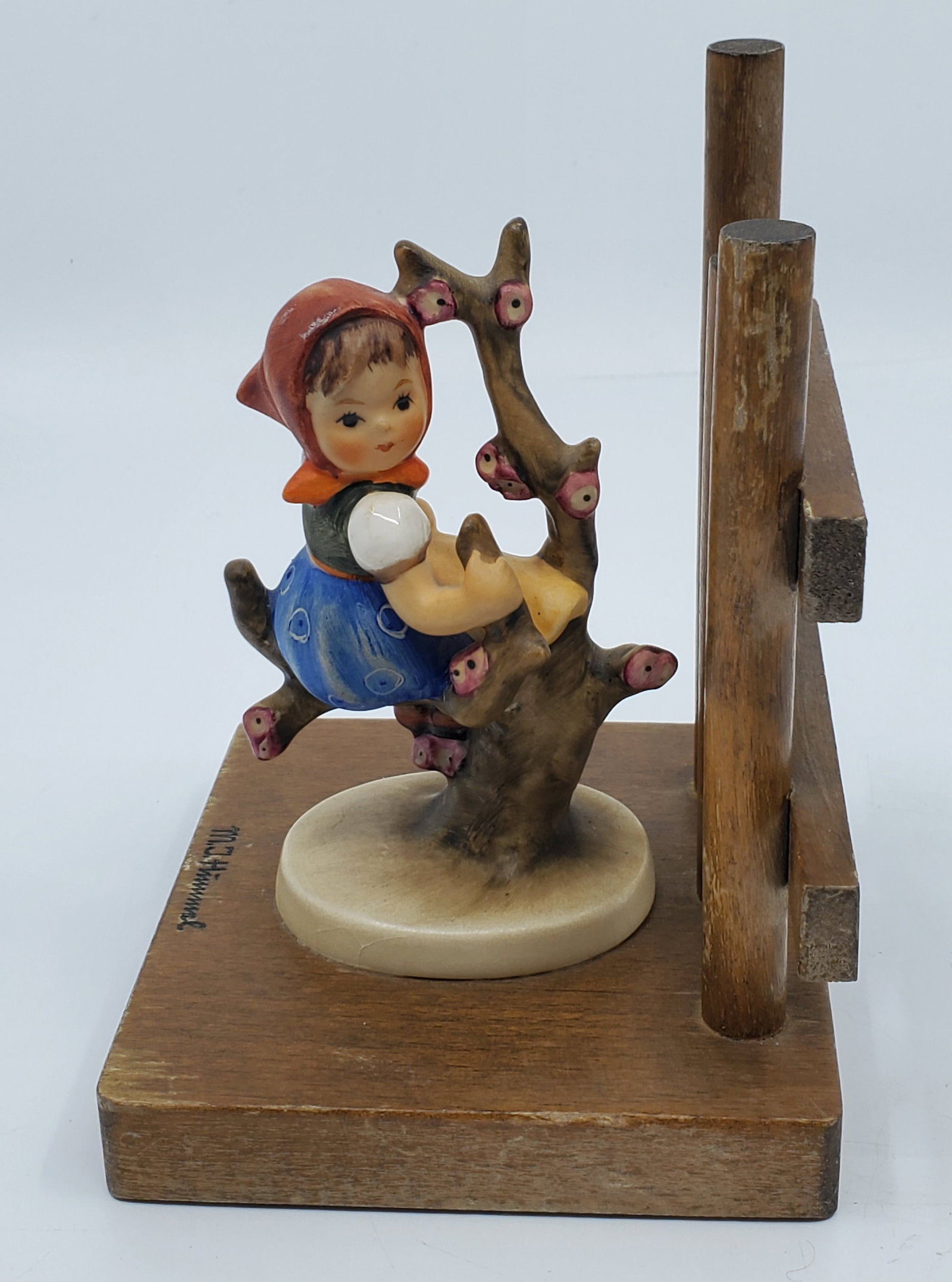 M. J. Hummel Western Germany Figurine on Sigmed Wood Base: 3 1/2 x 3 3/4" base, 5" high, Wood base signed with name on top front, logo on front edge, Western Germany on back edge, Figurine is fully intact, with some roughness on base front edge.