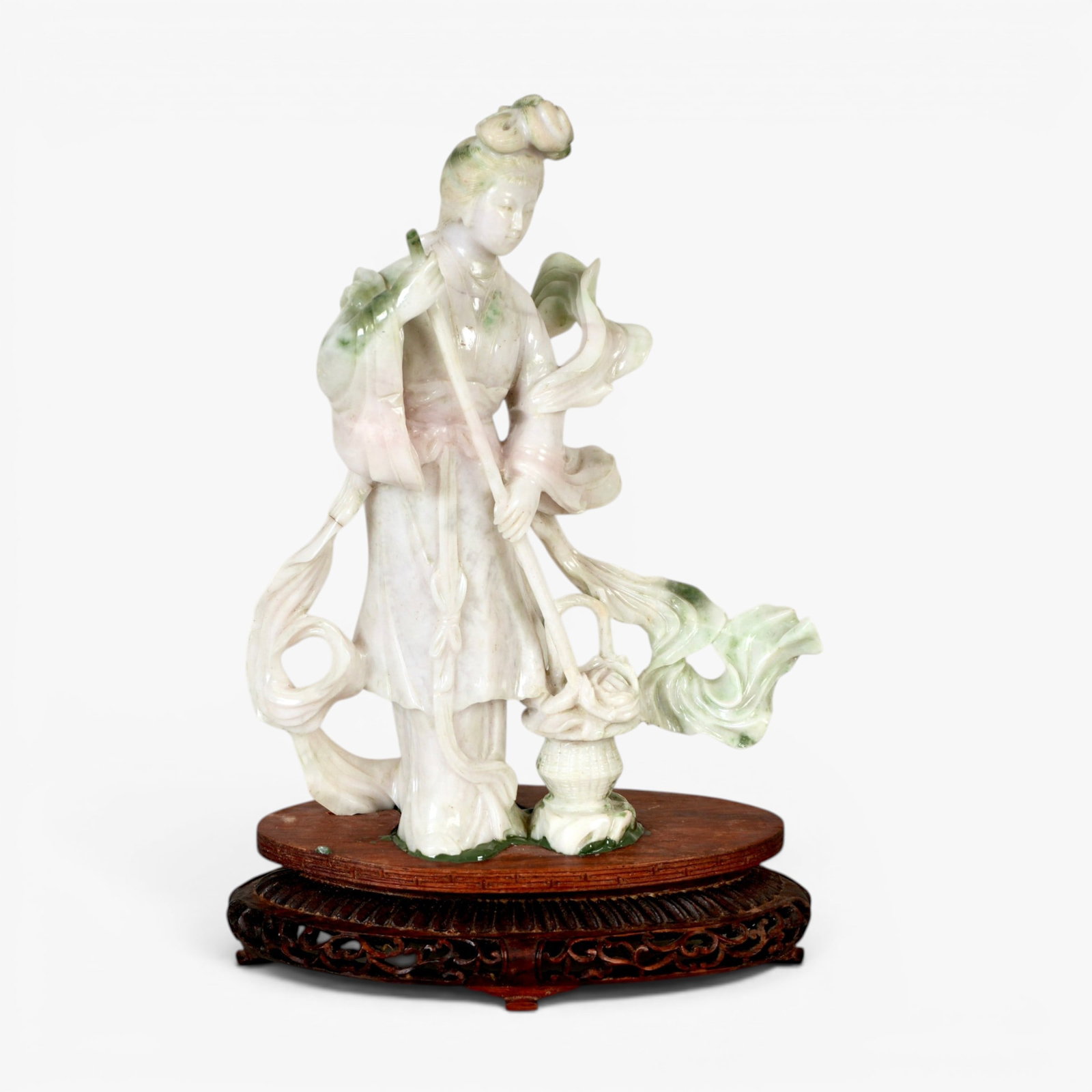 Chinese Jade Sculpture of a Maiden: 20th century, carved dressed in long robes with a billowing scarf over the shoulders, holding a staff above a basket of blossoming peony, the hair tied up in a high chignon and draping down the back, 