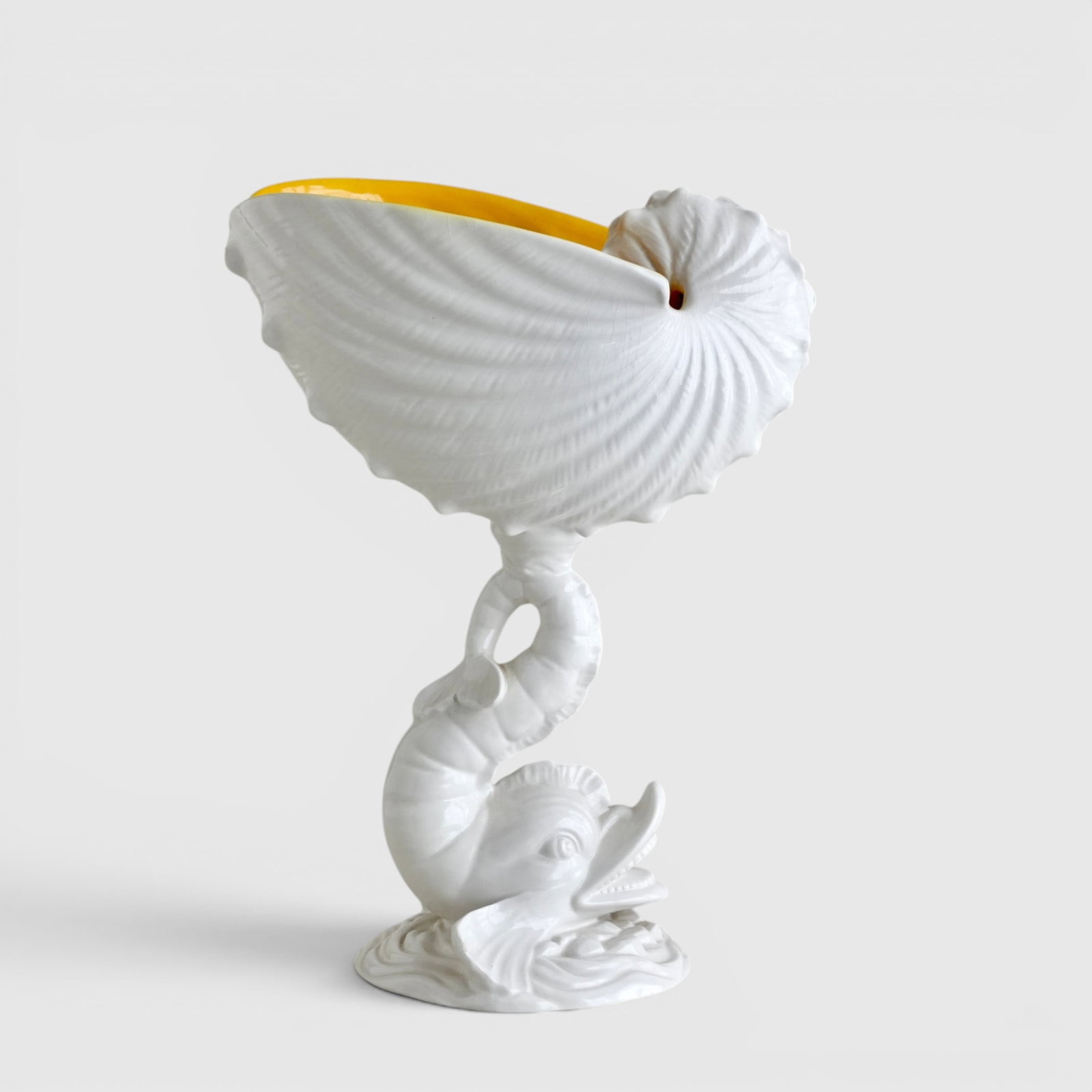 Italian White Porcelain Fish and Nautilus Shell Pedestal Dish: In the shape of a deep shell supported on a stylized dolphin stem and base, the interior glazed yellow, the exterior glazed white, marked on underside 'Italy' The collection of Frank and Irene Giuffri