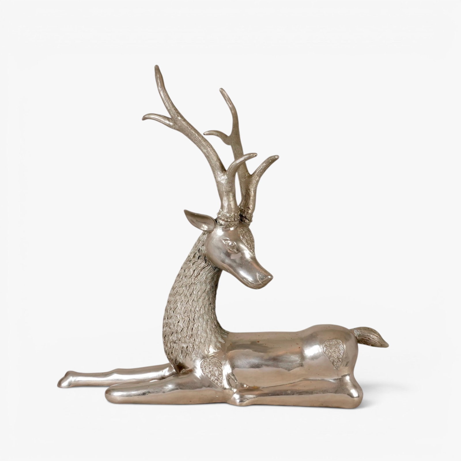 Indian Silvered Brass Deer: Resting in a recumbent position with one foreleg outstretched, the raised head with large antlers and a thick fur neck, adorned with raised medallions on the forehead, haunches and shoulders The