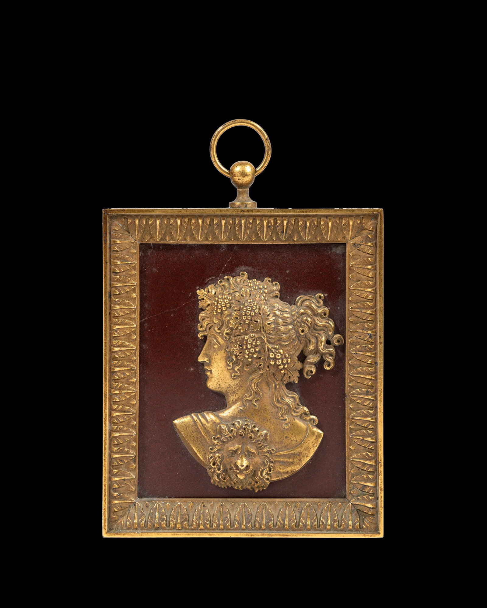 A GILT BRONZE PLAQUE, Rome, 19th century: h cm 11,8 x l cm 9.Depicting a female face on a red marble background, with a hairstyle adorned with bunches of grapes and vine shoots, draped in a cloak decorated with a lion motif. Signed on the rev