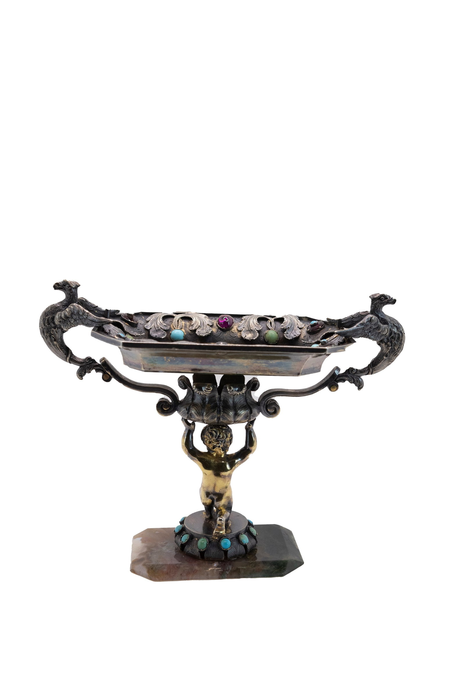 °A SILVER AND HARD STONE STAND, 20th century: h cm 11,5 x l cm 15 x p cm 6,9 Of octagonal form; central putto in vermeil; eagle-shaped handles; decorated with hard stones; resting on a faceted hard stone base.