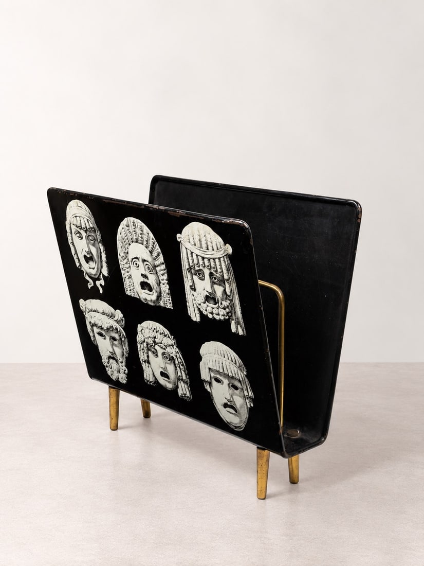 Piero Fornasetti (Milano 1913 - 1988), MAGAZINE RACK, circa 1960s: l cm 42,5 x h cm 39 Black lacquered metal with screen-printed decoration from the "Maschere" series, brass feet. Produced by Fornasetti, Milan.Catalogue notes: For the same model, with the same decora