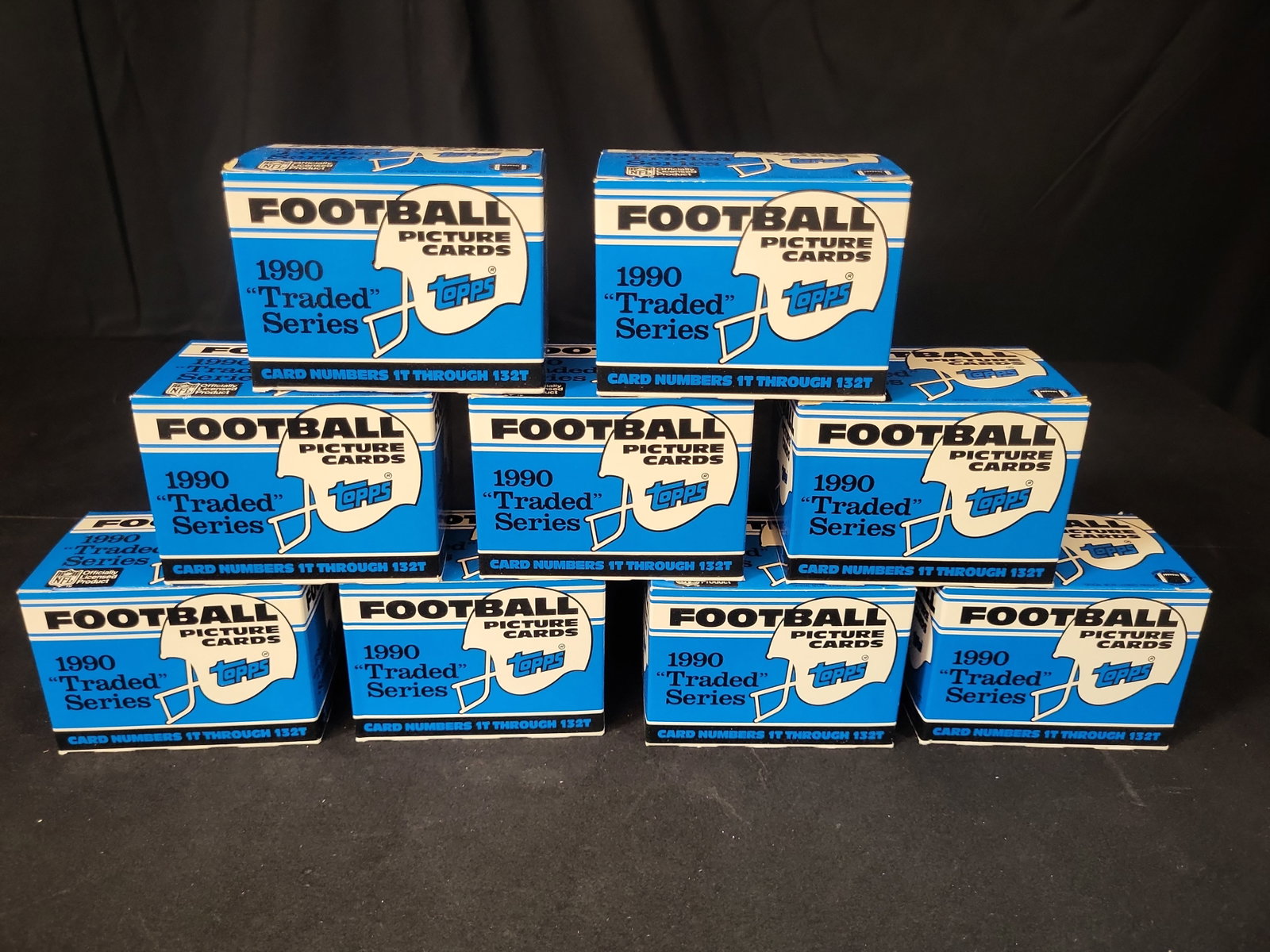 I03252 | Group of 9 1990 Topps Football Traded Series Factory Set Boxes: BID PER WHAT YOU SEE IN THE PHOTO, NOT THE AI GENERATED TITLES AND DESCRIPTIONS! This lot features a group of nine 1990 Topps Football Traded Series factory set boxes. Each factory box contains a comp