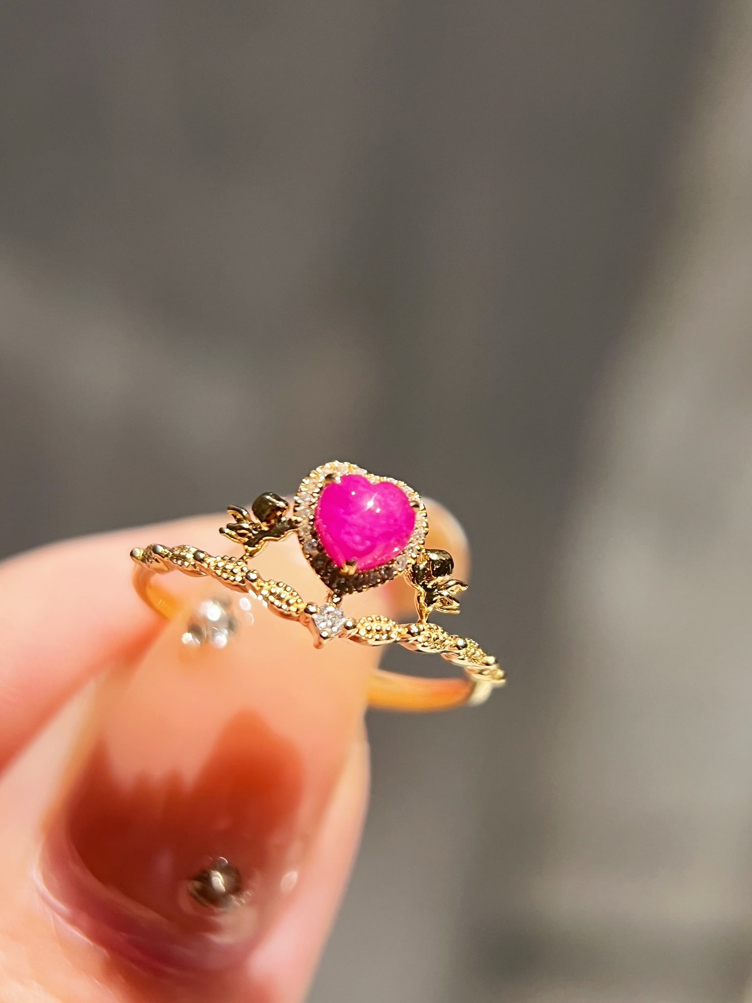 18K Ruby Cupid Ring: This ring features 18K gold, with a heart-shaped ruby centerpiece in rose hue, surrounded by a row of diamonds. The sides are designed with three-dimensional cupids, creating a crown-like appearance.