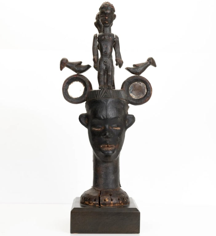 An African Shrine Head with Figures: Surmounted by a standing figure and birds, carved with recessed circular elements and mounted on a base. H:24in(60.9cm) W:10.5in(26.6cm)