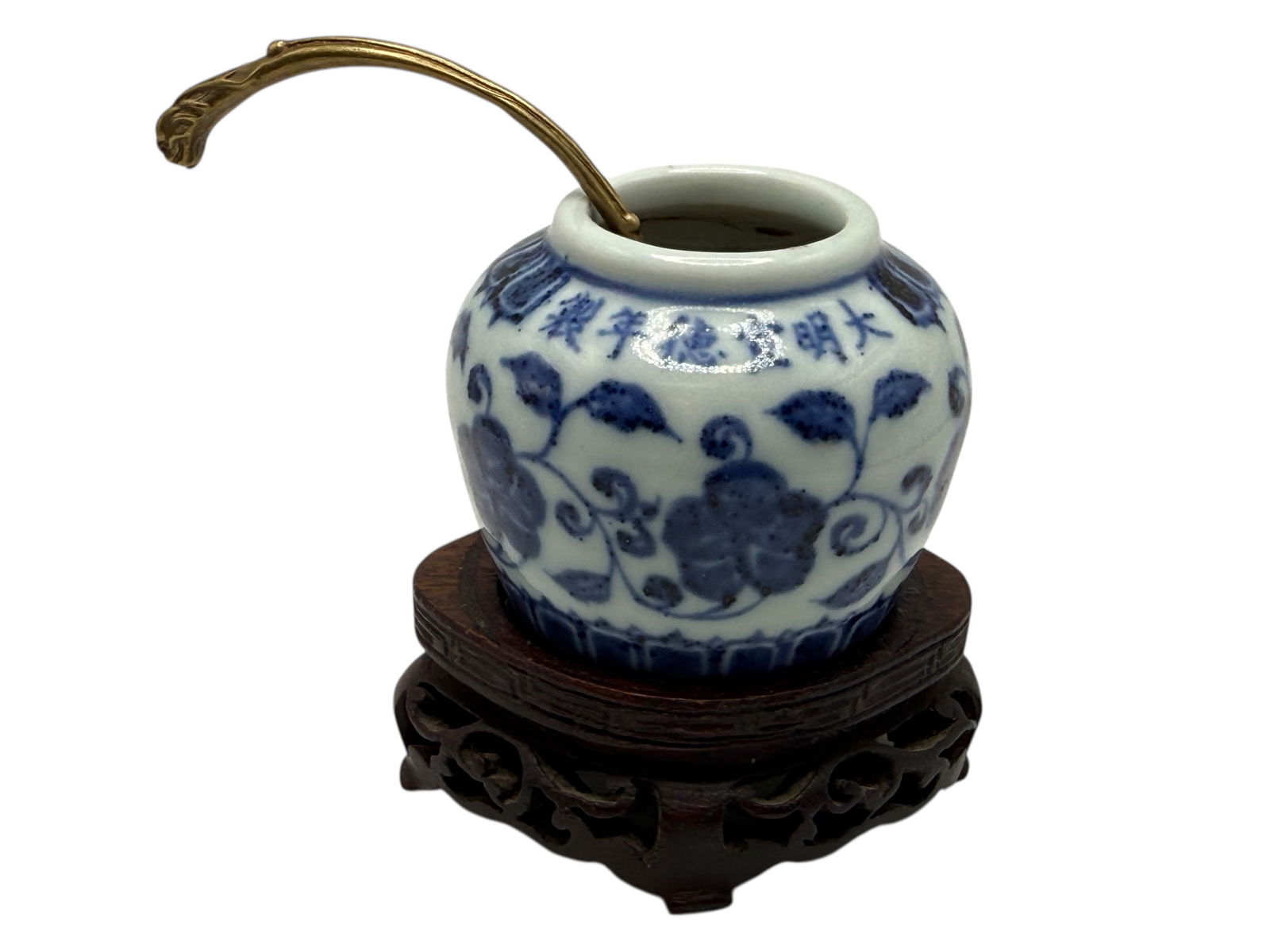 A Chinese Blue and White Jar with Floral Scrolls: The globular body painted with scrolling lotus beneath a band of characters around the shoulder. Mounted with a metal ladle and displayed on a carved wood stand. H: 2 inches
