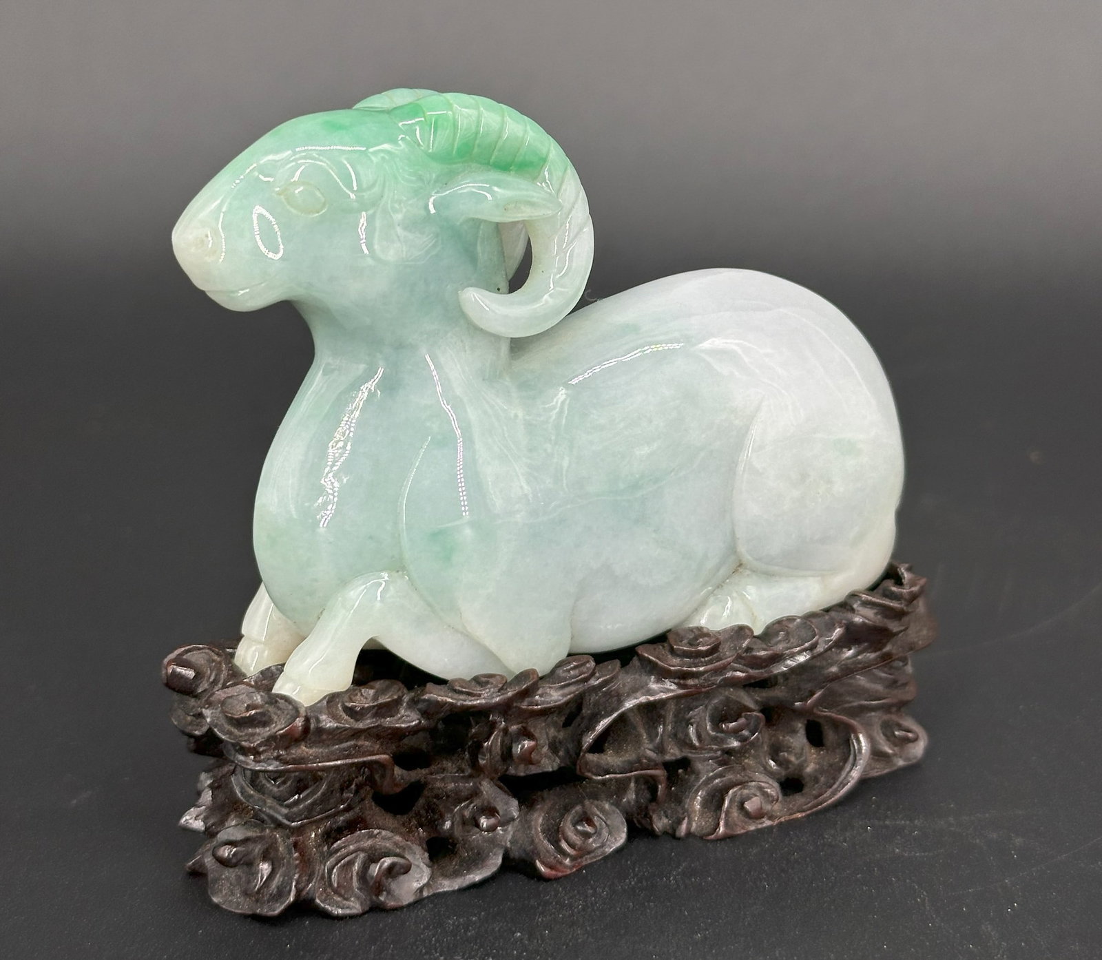 A Chinese Jadeite Figure of a Ram: Carved recumbent with curled horns, the stone of pale celadon tone with green inclusions, on a carved wood stand. W: 3 1/2 inches, D 1 1/8 inches, H: 2 1/2 inches (Excluding Wood stand)Wood Stand: H:1