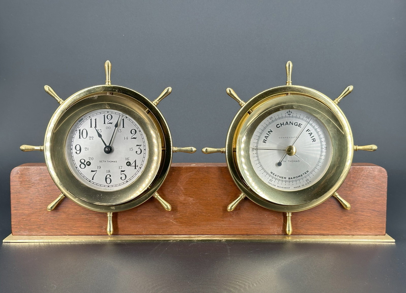 A Seth Thomas Brass and Wood Clock and Barometer S: Each circular brass case modeled as a ship’s wheel, mounted on a rectangular wood base. Size: H: 9 inches W:19 inches D: 3 inches
