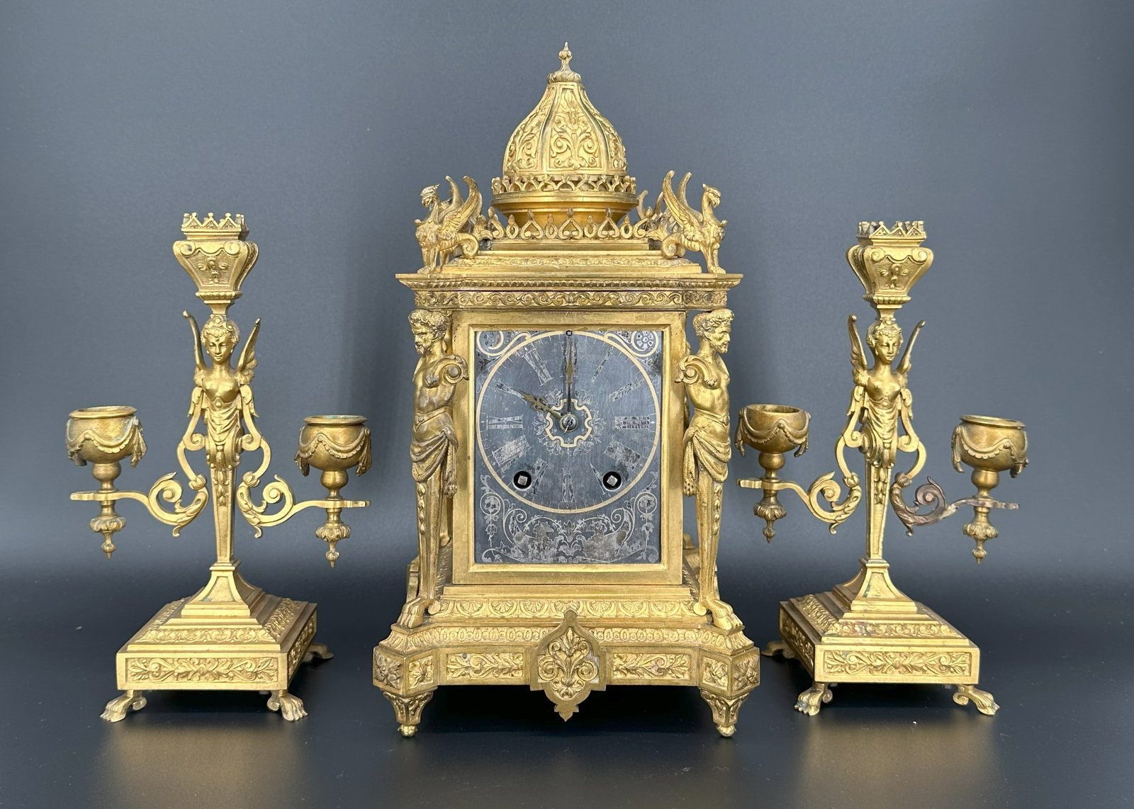 A French Gilt Bronze Figural Mantel Clock Garnitur: The architectural case flanked by classical figures and surmounted by a domed crest, with a pair of matching candelabra. Size : (Clock) H: 12 1/4" ; (Candle Holder) H: 9"