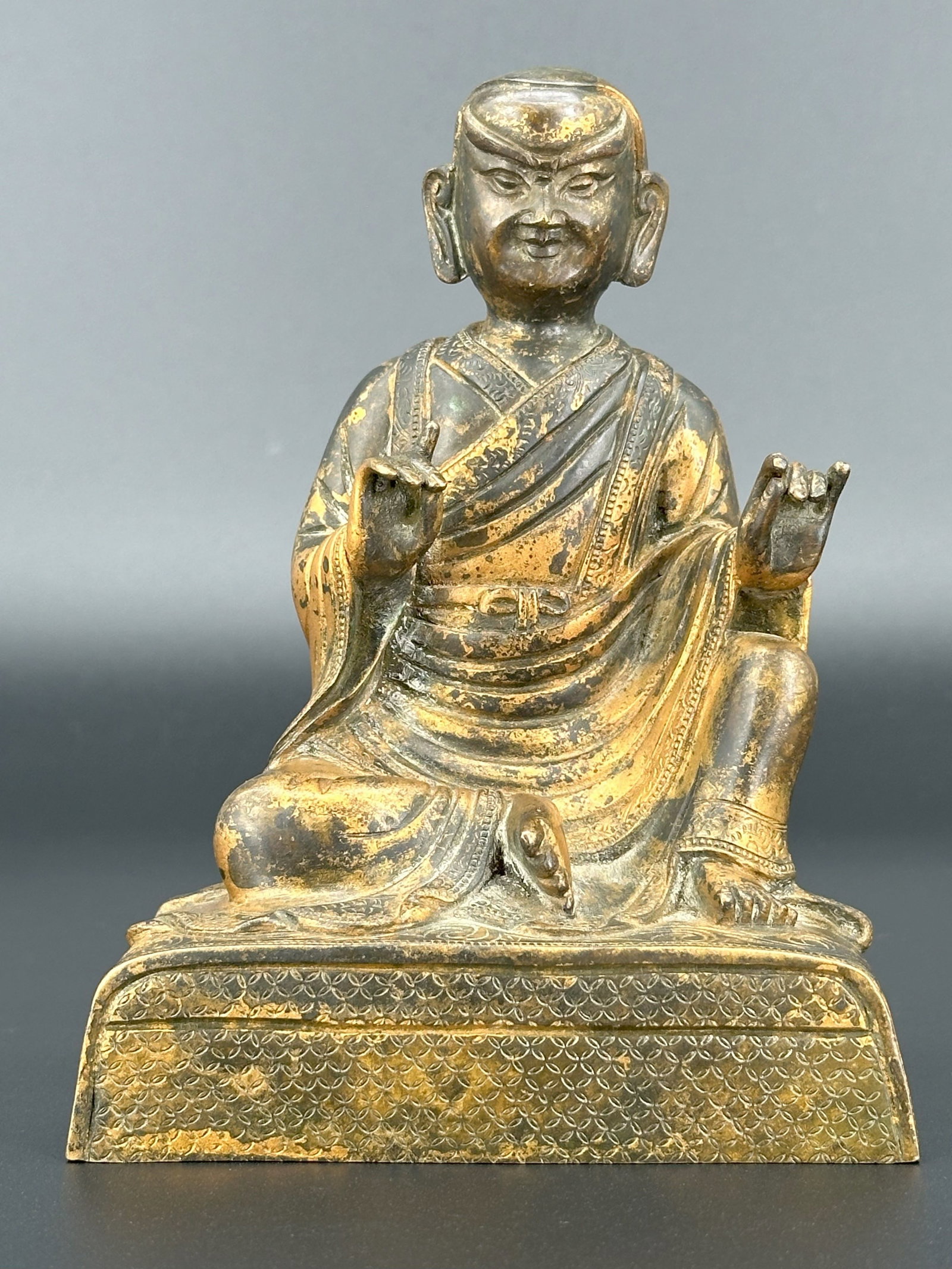 A Tibetan Gilt Bronze Figure of a Lama: Seated on a rectangular base, wearing layered robes and holding both hands raised in mudra. Size: H: 6 inches W: 4 1/4 inches D: 2 1/8 inches