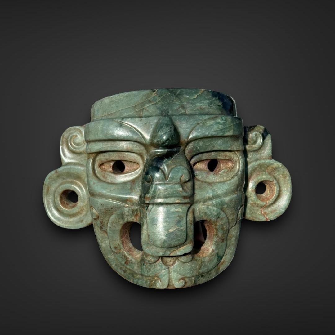 Pre-Columbian Maya jade deity mask: Guatemala, circa 600-900 CE. Maya jade deity mask intricately carved from a mottled green jade stone. The mask represents a deity, likely one associated with rulership, nature, or the underworld, give