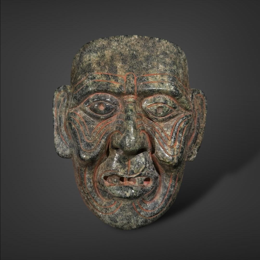 Pre-Columbian Maya god green stone mask: Mexico, circa 600-900 AD. Maya Huehueteotl mask, intricately carved from dark green stone, likely serpentine. Huehueteotl, the old fire god, is often depicted with aged, weathered features, reflecting
