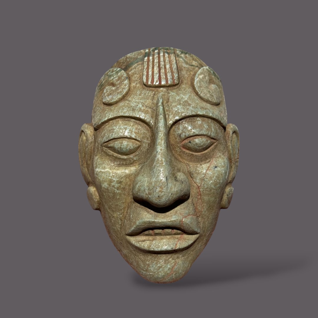 Pre-Columbian Mayan green stone mask: This Pre-Columbian Maya mask, carved from green stone, reflects the intricate craftsmanship and deep symbolic meaning associated with Maya ritual and ceremonial art. The face is finely sculpted with a