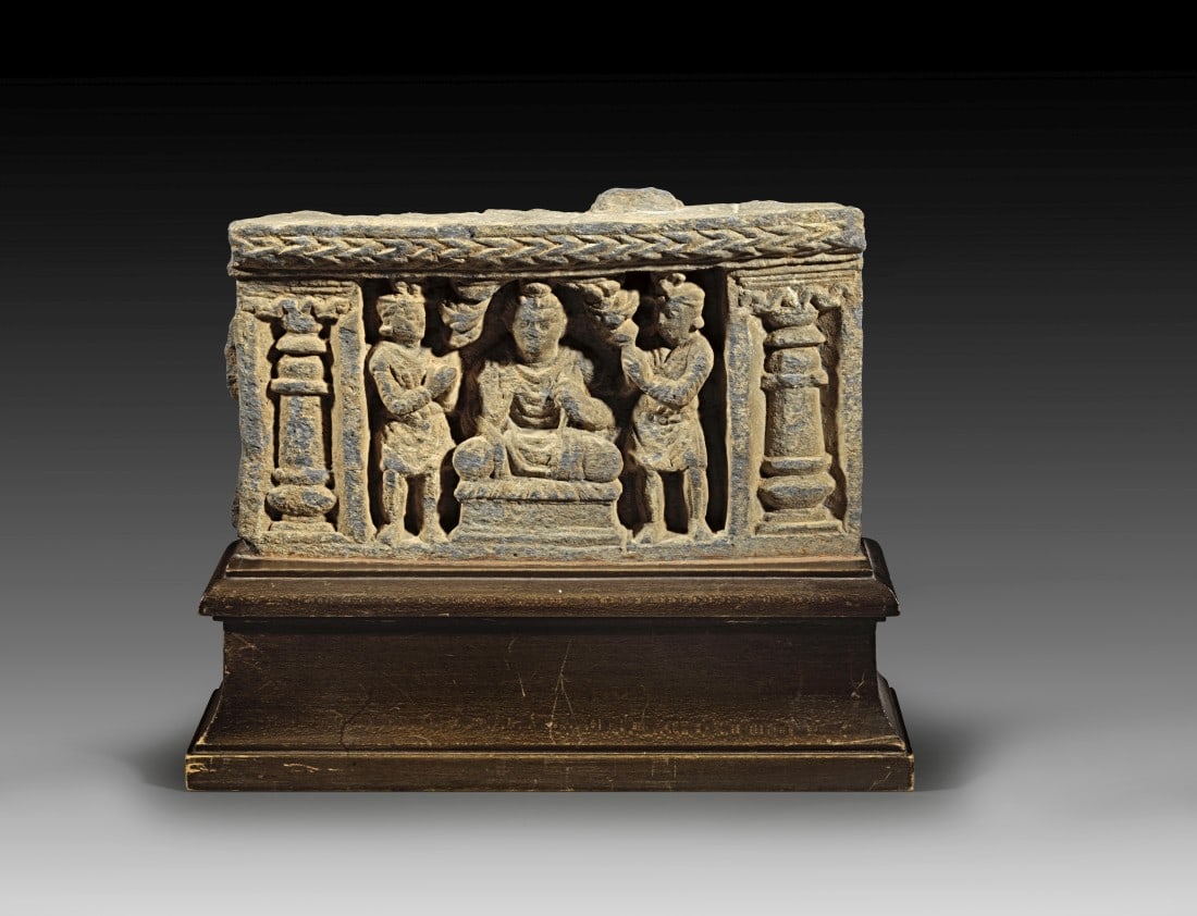Schistrelief mit Buddha.: Schist relief with Buddha, height 14.5cm. length 24.5cm, depth 3cm. Buddha is depicted in the center under a canopy of leaves in a meditation seat on a throne, his right hand in abhaya mudra. On both 