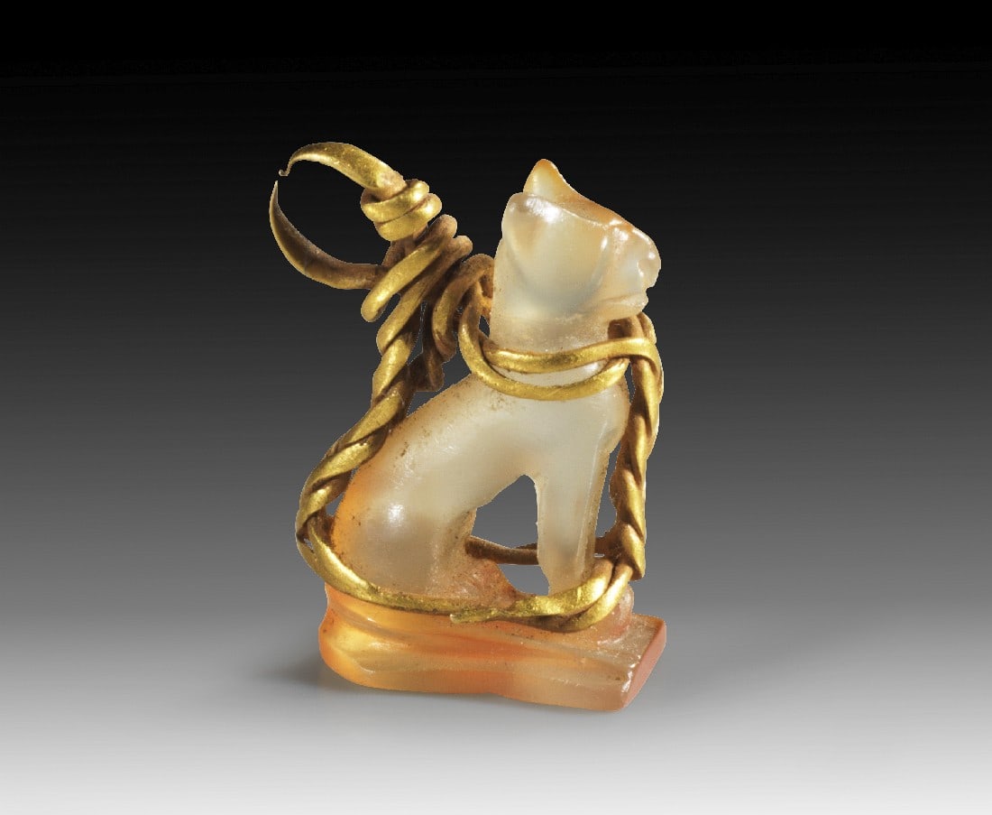 Anhänger in Form einer Miniatur-Katze.: Pendant in shape of a miniature cat. H 0.7cm, W 0.9cm, L 0.8cm. Carnelian and gold wire. Goddess Bastet in form of a sitting cat on a plinth, gold wire partially braided around the body, forming an ey