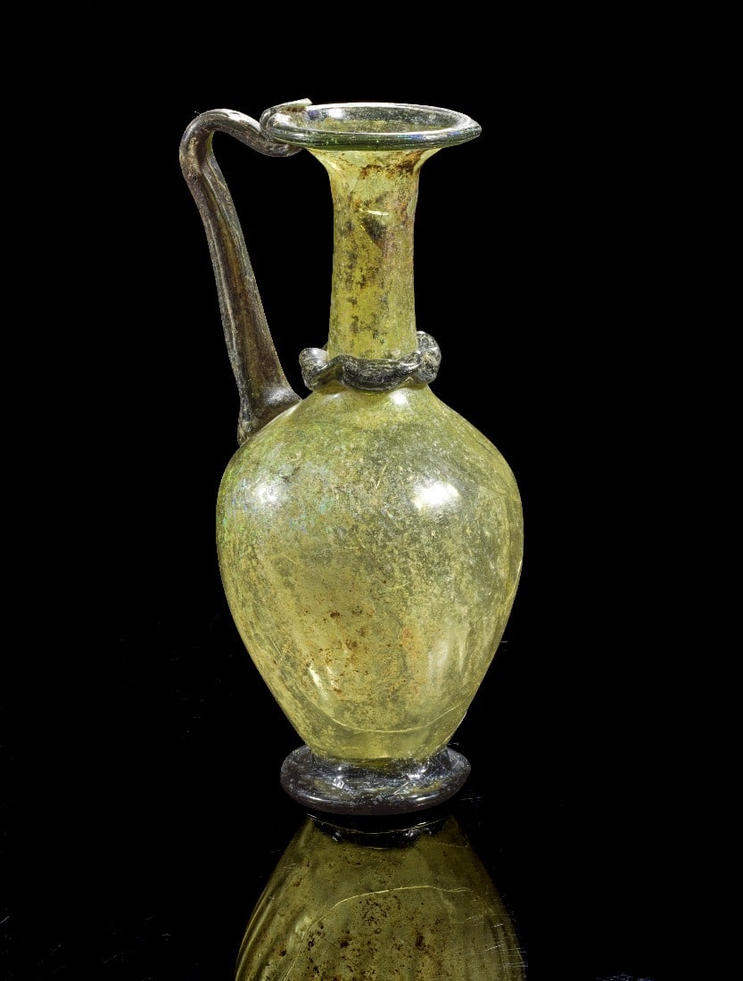 Kanne.: Jug. Eastern Mediterranean, 4th - 5th century AD. H 20,5cm. Green translucent glass. Globular body with tall neck. Foot, ring trail around neck, handle, and rim of opaque black glass. With invoice fro