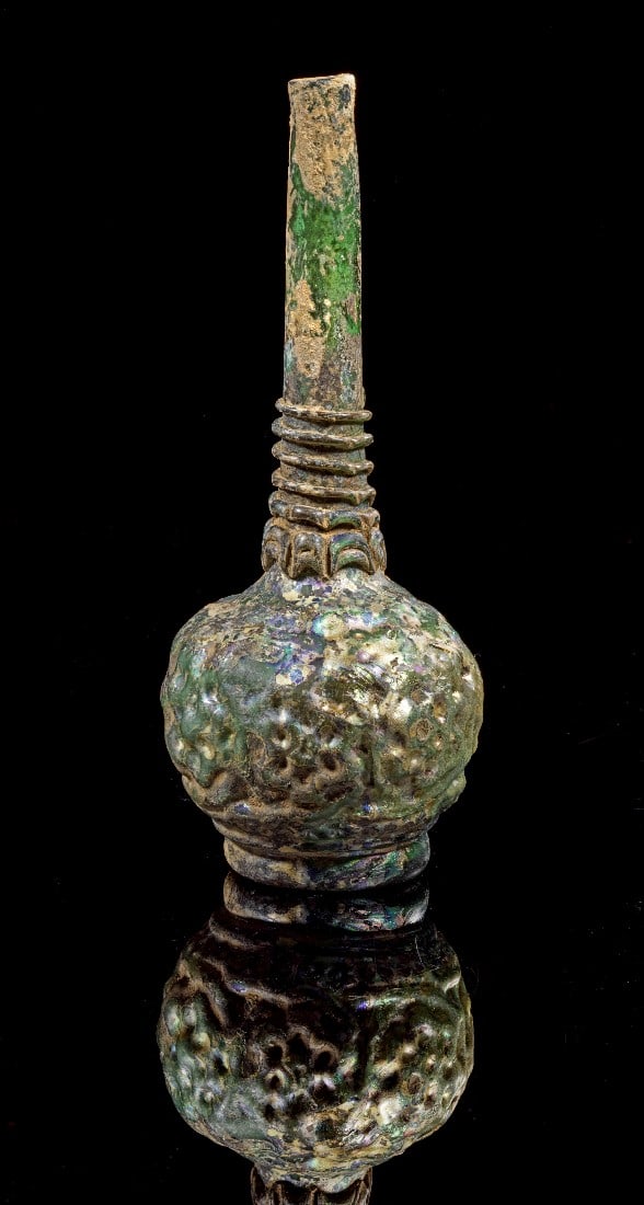 Formgeblasene Flasche.: Mold-shaped bottle. Islamic, 11th - 12th century. H 18,3cm. Green translucent glass. Globular body on thick ring foot, with tall cylindrical neck and flattened rim. Body with relief pattern depicting